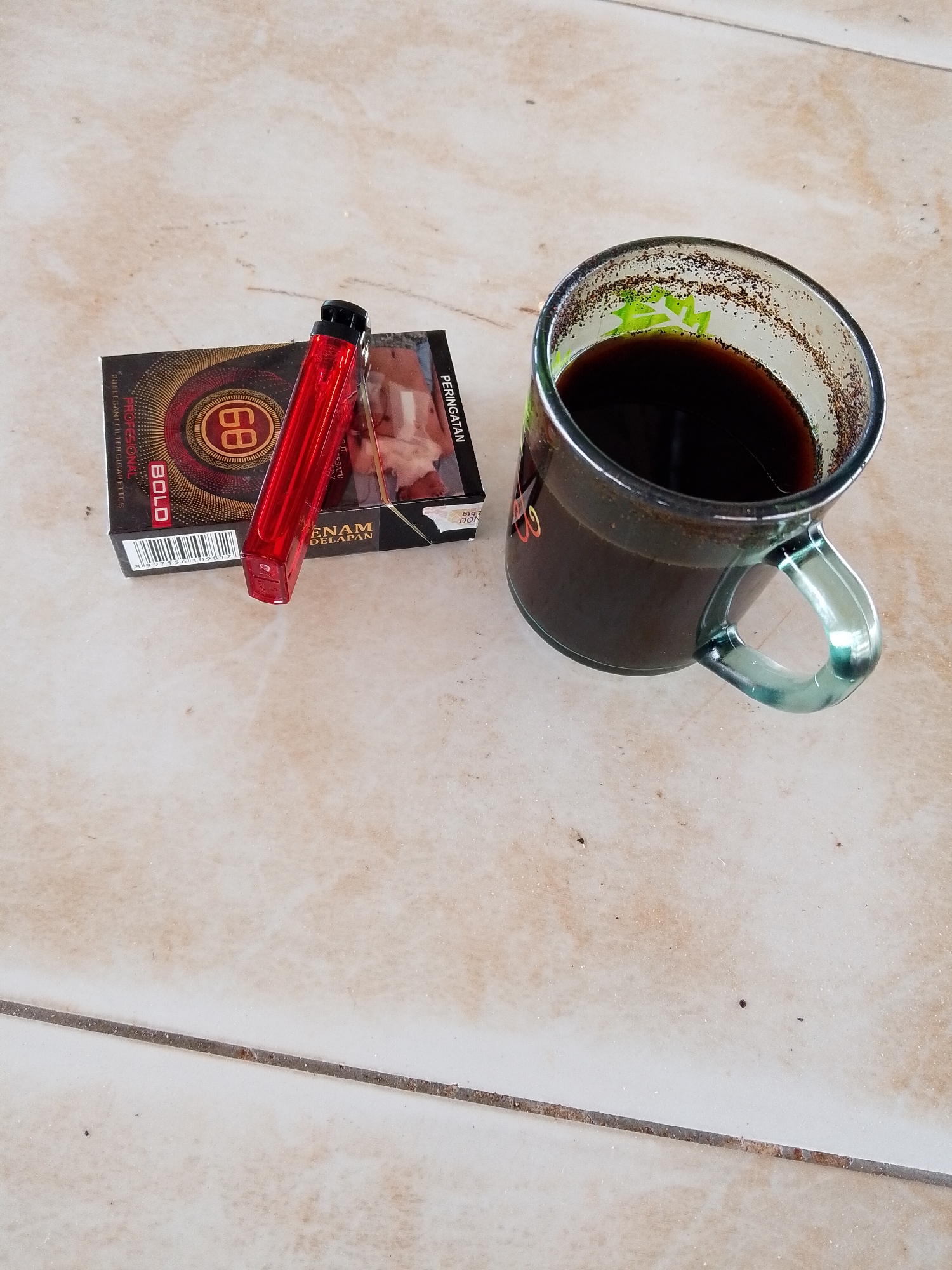 On a light-colored, tiled surface, likely an outdoor table, are three items: a pack of cigarettes, a red lighter, and a glass mug filled with a dark liquid, presumably coffee. The cigarette pack is branded "BOLD" and "68 ENAM DELAPAN." The lighter is simple and red. The mug is glass with a green tint and a clear handle. The coffee appears to have coffee grounds around the rim of the mug. The scene appears to be in Bukit Ketok, Indonesia, based on the cigarette brand being commonly found in that region. There are no people visible in the scene. The environment appears to be a casual outdoor setting.
