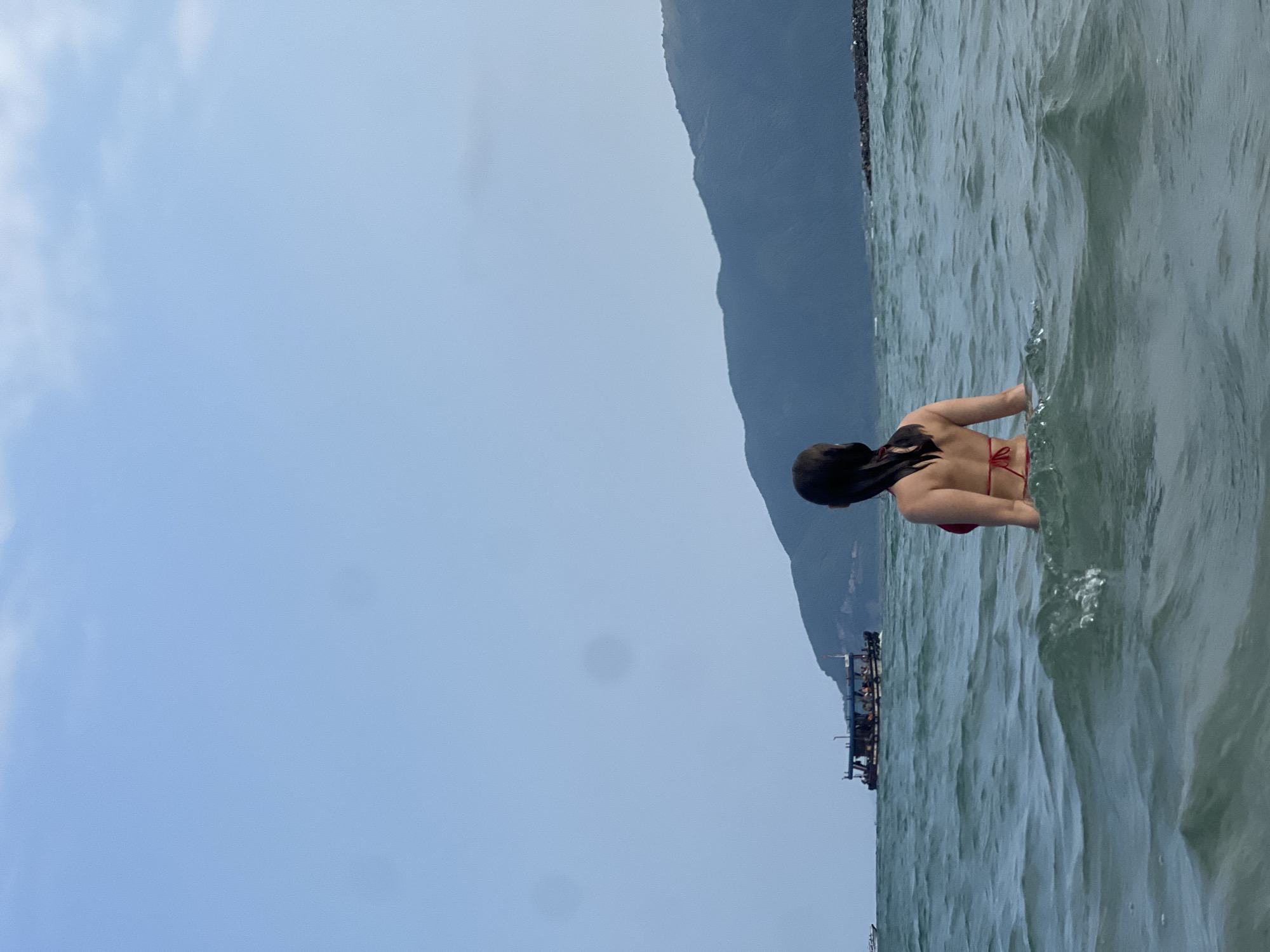 The image captures a serene coastal scene in Nam Nha Trang, Vietnam, featuring a person wading in the ocean. The main subject is a person, likely female, viewed from behind and waist-deep in the water. She has dark, wet hair and is wearing a red bikini top with straps tied at the back. Her gaze is directed towards the distant horizon and the mountainous landscape.
The setting is an open sea or large bay, characterized by calm, light blue-green water with gentle ripples and small waves. In the background, a majestic dark mountain range rises under a soft blue-grey sky, which appears bright but partly cloudy or overcast. Closer to the mountains, a long, dark boat or floating platform, with several indistinct figures visible on it, adds a touch of human activity to the expansive natural scenery.
The time of day appears to be daytime, likely late morning or early afternoon, given the ambient light. The atmosphere is peaceful, suggesting a moment of relaxation or leisure by the sea. No text is visible in the image.