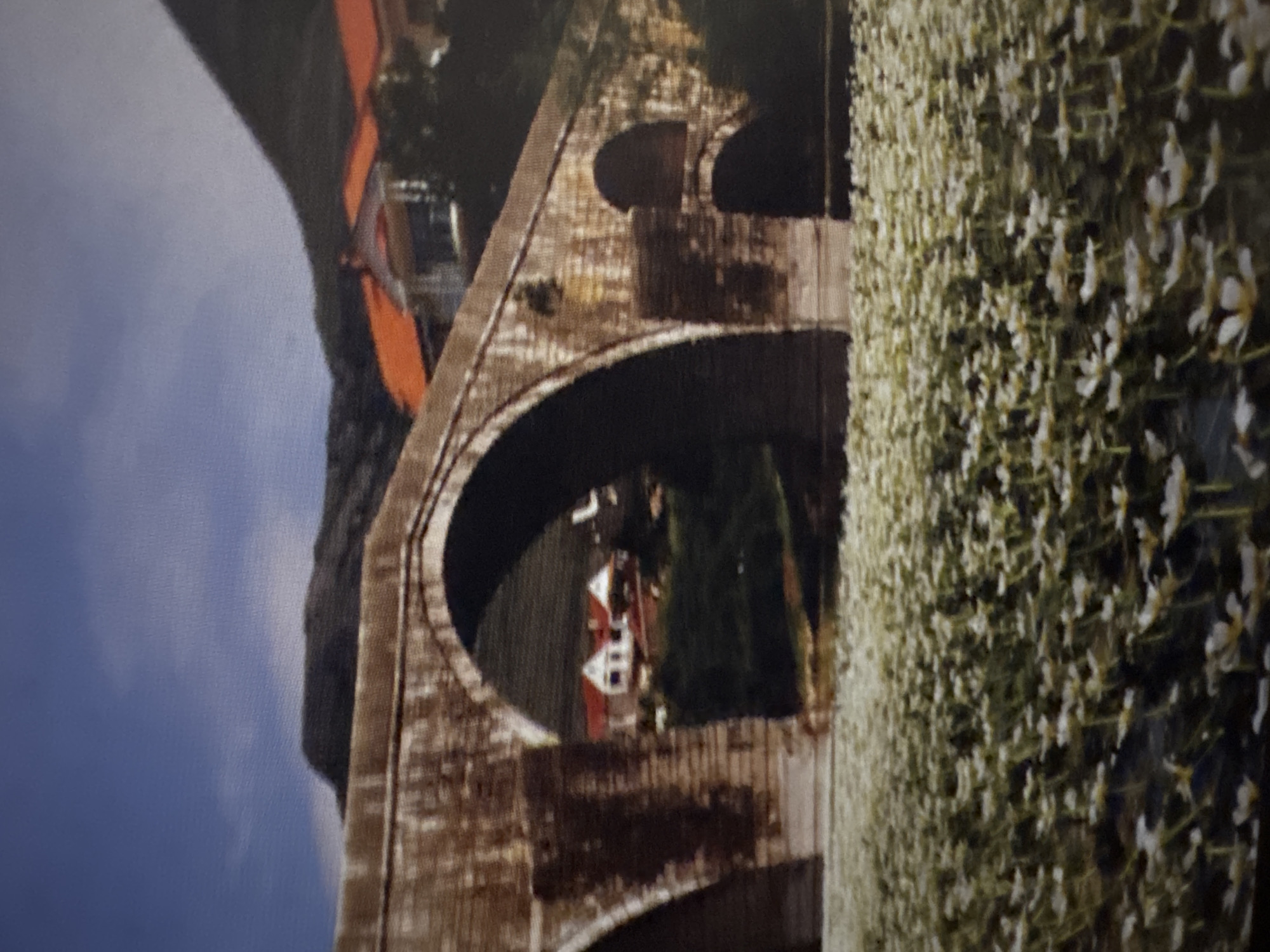The image displays an ancient multi-arch stone bridge traversing a body of water, with a dense field of small white flowers with yellow centers covering the water's surface in the foreground. The bridge, constructed from rough-hewn brown and gray stone, exhibits weathering and discoloration. Its main arch is prominent, flanked by at least two smaller rounded arches to its right and a partially visible arch to its left. In the mid-background, structures with reddish-orange tiled roofs are visible both on the right bank, partially obscured by a dark mountain, and through the left arch, where white-walled houses are nestled against a green hillside. A dark, rugged mountain range forms the distant background under a clear blue sky with minimal, wispy clouds. No individuals are depicted. The scene is located in the city of Unknown, Unknown.