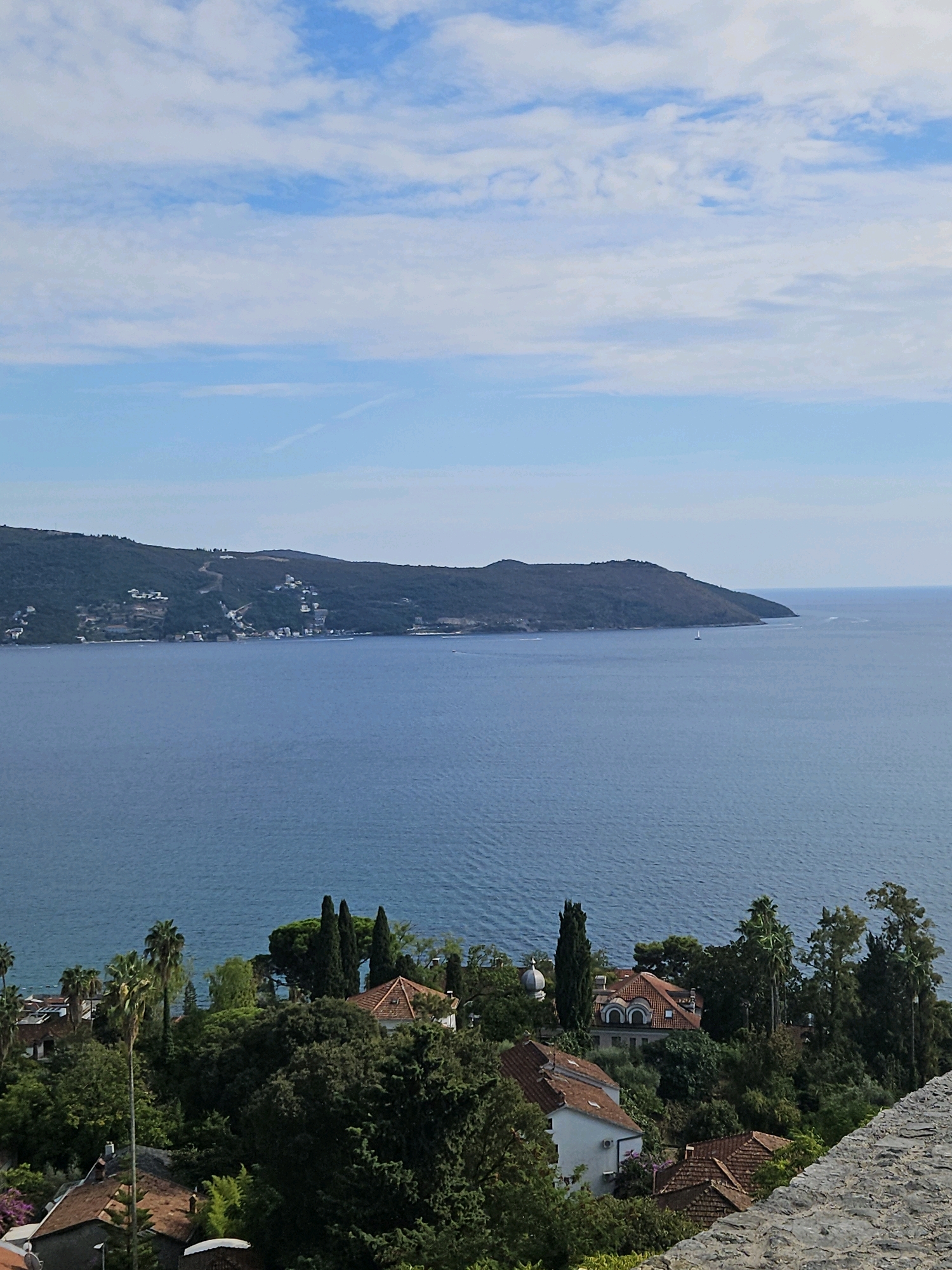 The image shows a panoramic view of the Bay of Kotor from a high vantage point in Herceg Novi, Montenegro. The foreground features a stone wall, with dense green foliage and red-tiled roofs of houses nestled amongst the trees. The houses are characteristic of Mediterranean architecture, with some featuring white walls and orange-tiled roofs. Further out, the deep blue water of the bay stretches towards a verdant, hilly coastline dotted with buildings. In the distance, the coastline curves and disappears into the horizon under a bright blue sky with scattered white clouds. A lone sailboat is visible on the water. The scene conveys a sense of tranquility and natural beauty.