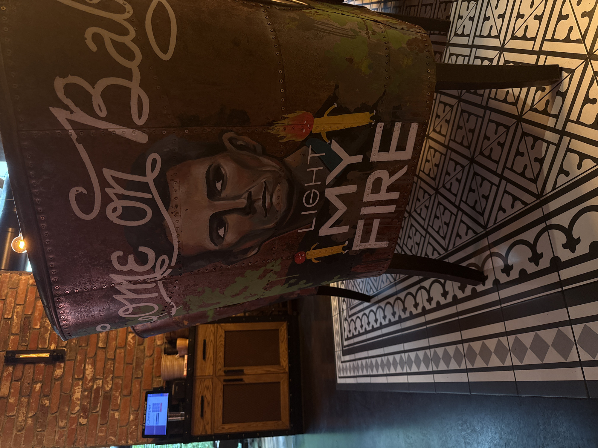 The image depicts the interior of a commercial establishment, identified as Bar "Jim," located at Ulitsa Goroda Izhevska, 252, Izhevsk, Russia. The central focus is the backrest of a chair, likely a bar stool or lounge chair, adorned with custom artwork. The artwork features a portrait of Jim Morrison, alongside the partial inscription "Come on Baby" and the prominent text "LIGHT MY FIRE," accompanied by two cartoonish burning matchstick figures. The chair's surface is metallic and riveted, displaying a distressed finish with brown and green hues.
To the left of the chair, a red brick wall is visible, upon which a vertical black sconce with a glowing filament bulb is mounted. Beneath the sconce, a wooden cabinet featuring two drawers and two mesh-fronted doors is positioned. On the cabinet's top surface, a monitor displaying a blue screen with "Keeper" visible (indicative of a POS system), a stack of white plates, and several yellow cups or small bowls are arranged. An additional bare Edison-style bulb light fixture is partially visible at the top right, contributing to the warm, subdued lighting of the space. The floor consists of plain dark tiles on the left and an ornate pattern of black and white decorative tiles, featuring geometric stripes, diamond patterns, and curvilinear designs, beneath the chair's legs. No individuals, actions, or interactions are present in the scene, which exhibits a static, industrially-inspired, and retro ambiance.