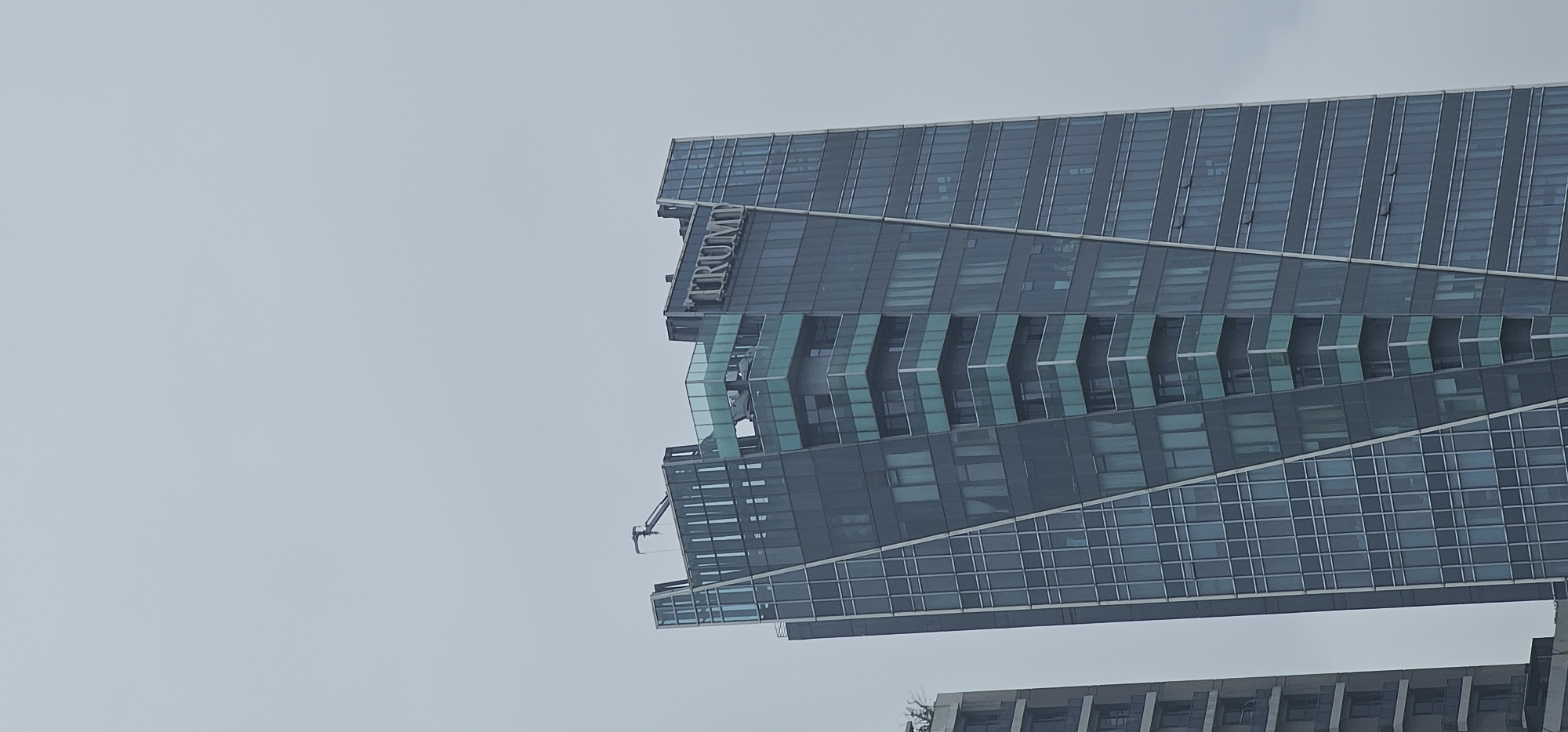 A wide-angle, low-angle, vertical shot captures the upper section of a modern skyscraper against a hazy, overcast sky. The building, identified by the prominent "TRUMP" lettering in silver on its facade, is designed with a striking architectural style. Its exterior is predominantly glass, with a dark, reflective blue hue, segmented by angular white and silver lines that create a dynamic, almost twisting appearance.
The building features several protruding sections with light teal-tinted glass balconies that step inward as they ascend, creating a tiered effect. On the very top of the building, a small crane-like structure is visible. To the left of the main skyscraper, another building of similar height and modern design is partially visible; it is also clad in dark glass with horizontal window bands.
The scene is devoid of any individuals or discernible activity, presenting a static architectural study. The overall atmosphere is muted due to the overcast weather. The building is located in Makati, Philippines.