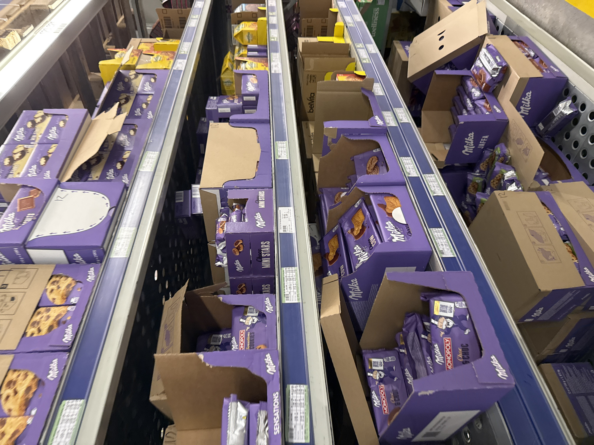 The image depicts shelves stocked with Milka brand chocolate products in what appears to be a retail environment, possibly a supermarket or grocery store. The scene focuses on three shelving units, with Milka items in various box and package sizes. The boxes and products are purple and brown. The items include "Mini Stars," "Monopoly," and "Jaffa" flavors, along with "Sensations" cookies. Some boxes are open or slightly damaged. Other items such as "BelVita" and "wellness" products can be seen on shelves behind the Milka displays. The location, Stara Zagora, Bulgaria, is not discernible from the image content alone, though it might be determined from other sources if available.