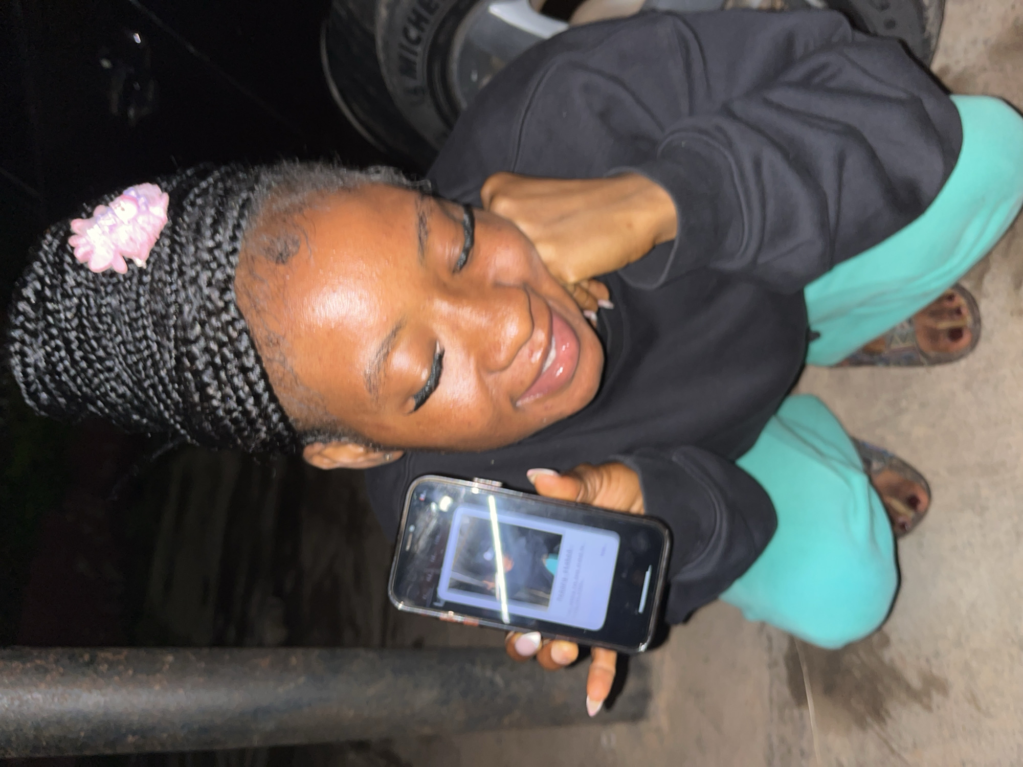 A young Black woman with braided hair adorned with a pink flower is crouching outdoors, holding up a smartphone. She is wearing a black sweatshirt and teal sweatpants, with sandals on her feet. The phone displays a polaroid-style image. In the background, a metal pole, a car tire with "MICHELIN" visible, and a dark, unpaved surface suggest an outdoor, possibly urban or semi-urban environment. The lighting indicates it is likely nighttime. The location is the city of Jos, Nigeria.