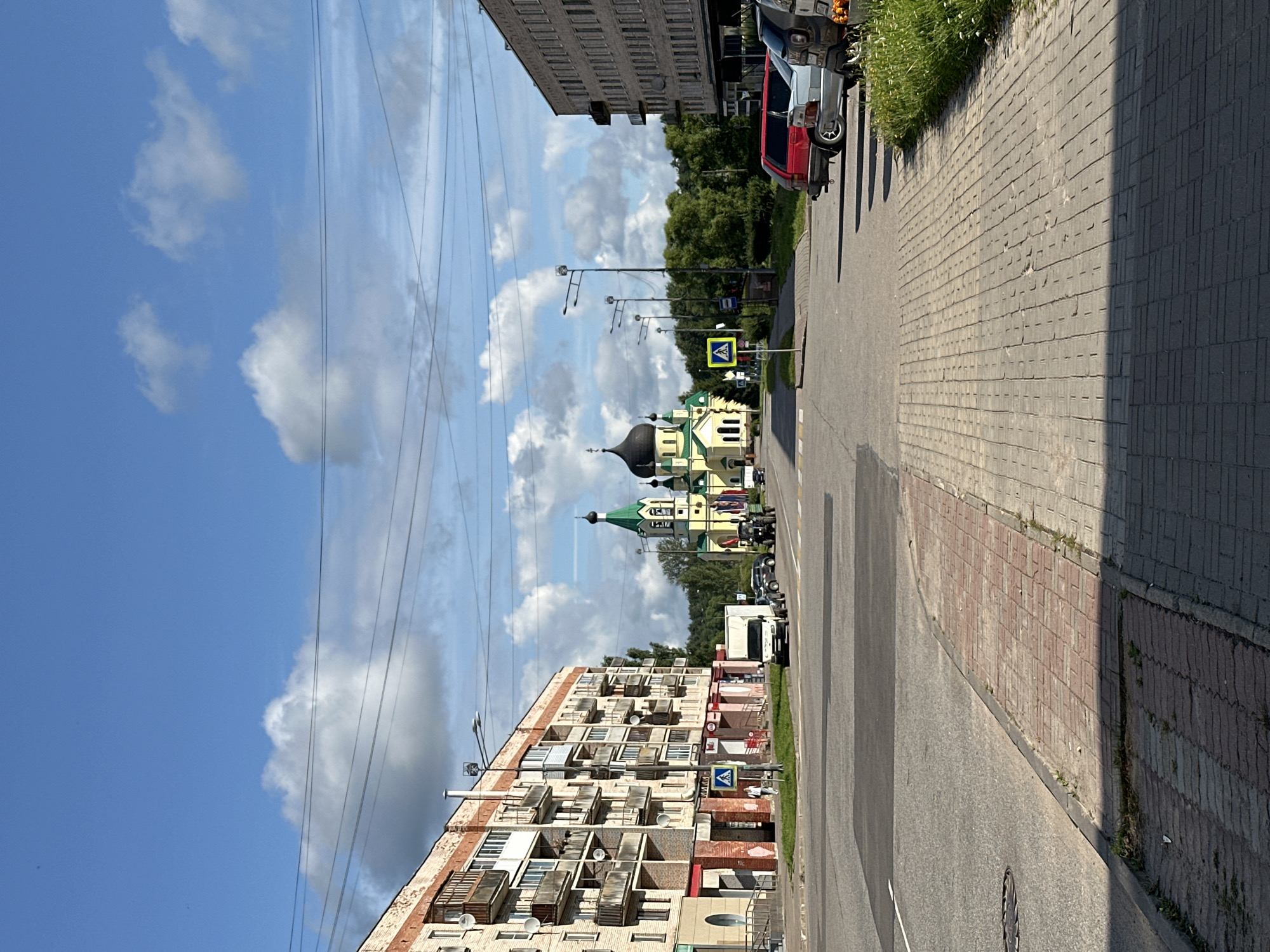 The image depicts a street scene in Slantsy, Russia, on a bright, partly cloudy day. A multi-story brick apartment building, featuring numerous balconies, lines the left side of the street. On the right, a taller, more modern brick building is visible, with parked cars in front of it. In the background, a prominent green and yellow church with a dark dome stands prominently, surrounded by trees. The street itself is paved, with a pedestrian crossing in the foreground. Several vehicles, including a white truck, cars, and a military-style vehicle, are present on the street. Power lines crisscross the sky, and streetlights line the sidewalks. The overall environment suggests an urban setting with a mix of residential and religious architecture.