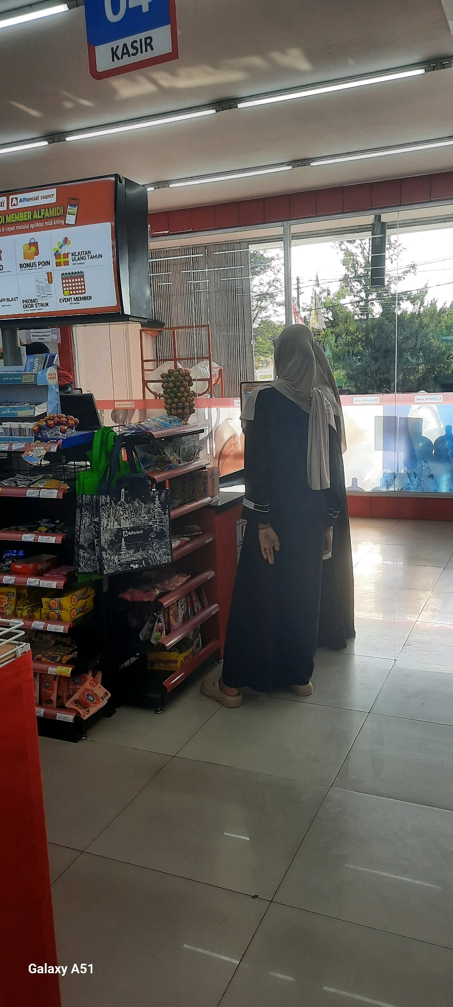 The image shows the interior of an Alfamidi minimarket in Wlingi, Indonesia. Two women, both wearing hijabs and long dresses, are standing near the checkout counter. The woman on the right is partially visible from the back, while the woman on the left is facing away from the camera and towards the counter. In front of them, the checkout counter area displays various products and promotional materials. A shelf unit to the left is stocked with packaged goods, including snacks and beverages. Above the counter, a sign indicates "KASIR" (cashier) with the number "04". The floor is tiled with light-colored ceramic tiles. The background reveals a large glass window with a view of trees and the sky outside. The overall scene captures a typical transaction or shopping experience within a convenience store.