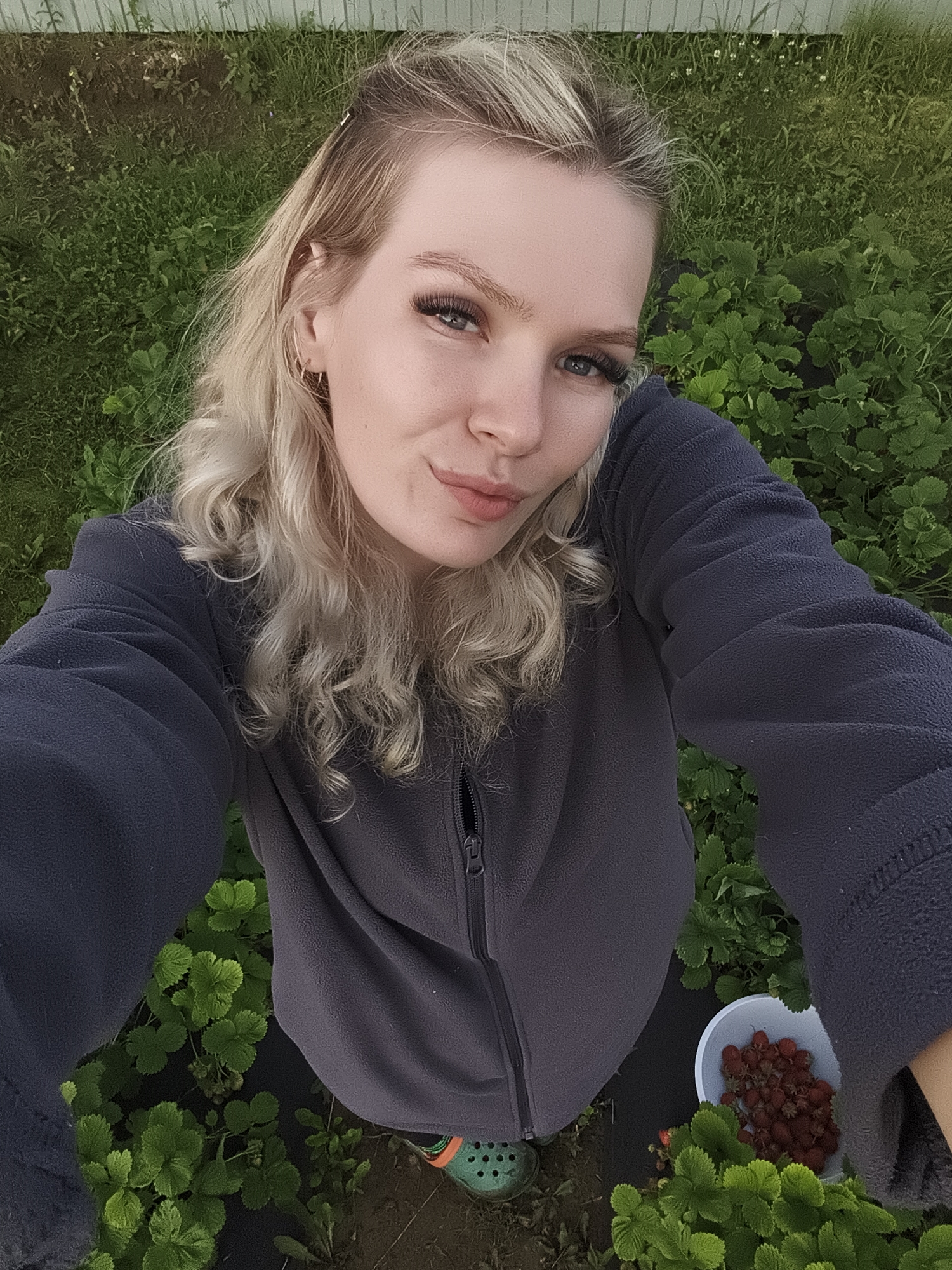A young woman with blonde curly hair is taking a selfie in a garden with strawberry plants. She is wearing a dark grey fleece jacket and green crocs. A small white bucket filled with strawberries is visible next to her. The background consists of green foliage and a white fence. The image appears to have been taken in Русский Вожой, Russia.