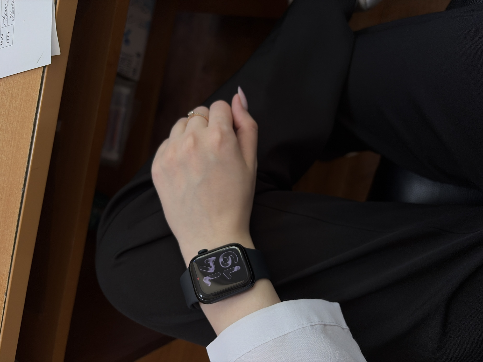 A medium shot depicts a person seated, presumably indoors, with their legs crossed. They are wearing black trousers and a white shirt with a small part of the sleeve visible. On their left wrist, they are wearing a black smartwatch. The time displayed on the watch face appears to be 17:16. The person is also wearing a ring on their left hand. To the left of the frame is a wooden desk or table with a piece of paper on it, displaying text that could be scheduling information with times like "18:30" and "19:00," possibly referring to the names "Alexei" and "Medvedev." The location may be Chaikovsky, Russia, although this cannot be definitively determined from the image alone.