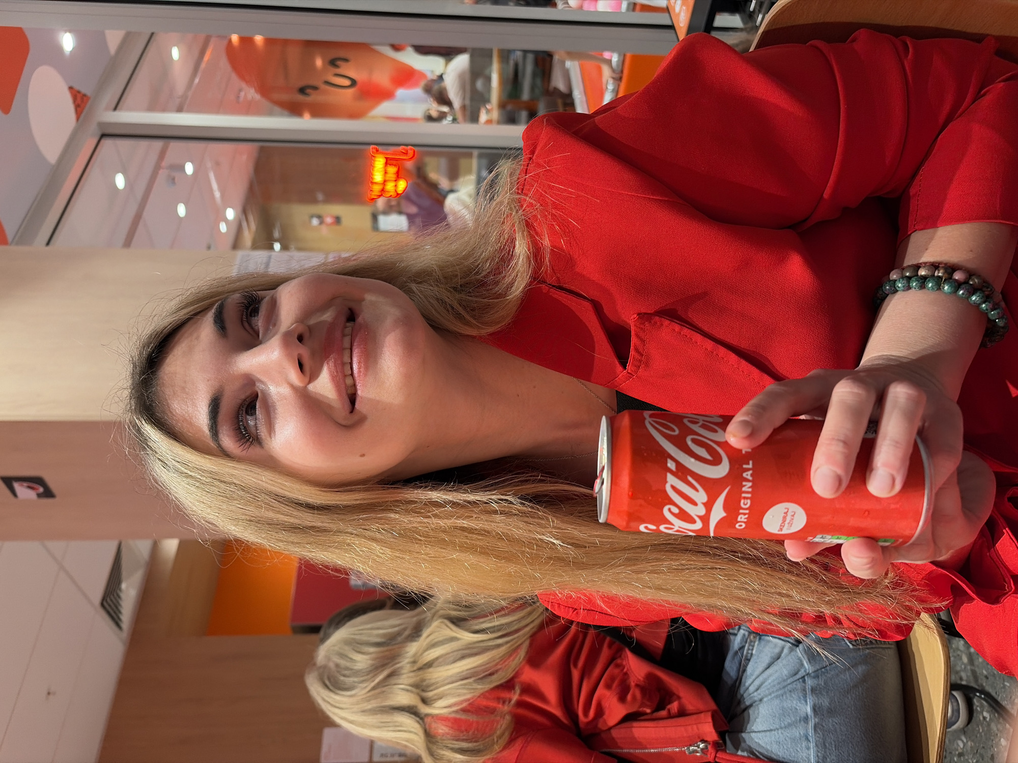 An indoor, eye-level portrait shot captures a young woman with long blonde hair, dressed in a bright red top or blazer, seated at what appears to be a table in a bright commercial space, possibly a restaurant or cafe in Budva, Montenegro. The woman is smiling broadly, looking slightly upwards and to her left, with a pleasant expression. She holds a red Coca-Cola Original can in her right hand, with her fingers wrapped around it. Two beaded bracelets, one green/dark and one multi-colored with green and pinkish beads, are visible on her left wrist.
The background shows reflections through a large glass window or partition, indicating an adjacent area or street scene. Visible in the reflection are elements like white lights on a ceiling, stylized orange and white graphic patterns on a wall, and an illuminated sign that appears to be yellow or orange, with part of a letter resembling an "F" or "E" visible. Another person, possibly with blonde hair, is partially visible in the lower right background, also wearing a red jacket and light blue jeans, suggesting a companion or another patron in the venue. The setting appears well-lit, suggesting daytime.
Text visible on the can clearly reads "Coca-Cola ORIGINAL." Below "ORIGINAL," there is some smaller text partially visible, which seems to be in a foreign language, possibly "SZUKAJ TRZWI" or similar.