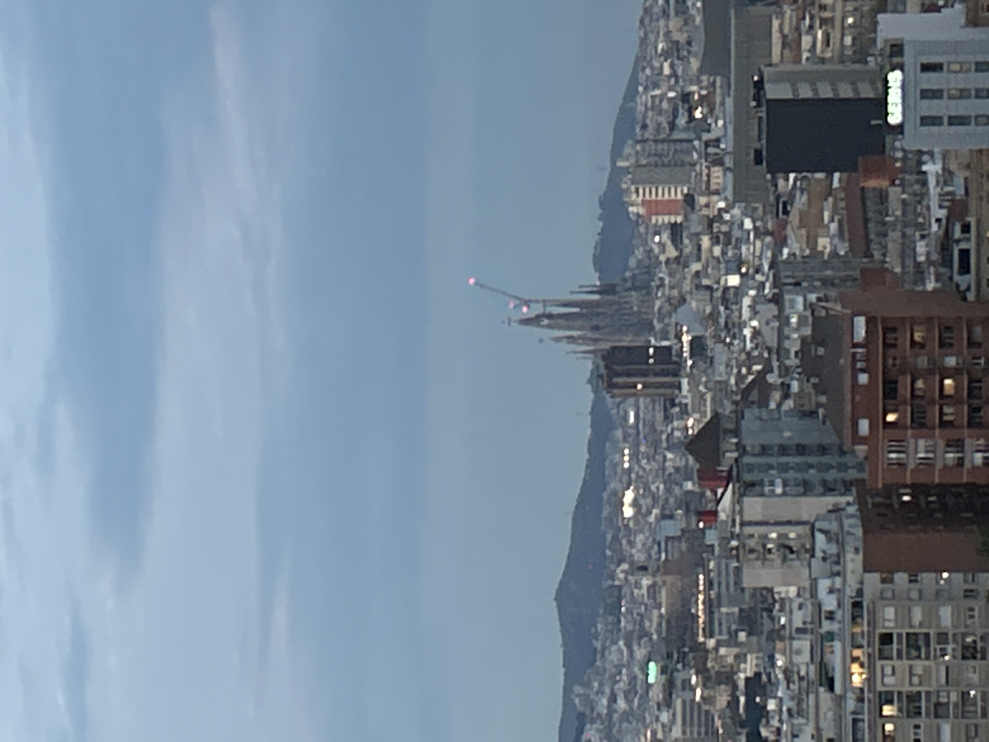 This is an elevated, panoramic view of the city of Barcelona, Spain, captured during twilight or dusk. The image depicts a vast urban landscape stretching towards a distant mountain range under a soft, light blue-grey sky that occupies the upper half of the frame.
The most prominent subject is the Sagrada Familia basilica, identifiable by its distinctive spires, which stands tall amidst the surrounding buildings in the mid-ground, slightly to the left of the center. Construction cranes, equipped with red warning lights, are visible atop its highest points, indicating ongoing work.
The city itself is densely built, characterized by a mix of residential and commercial buildings of various heights and architectural styles. Many buildings feature illuminated windows, and scattered streetlights or building lights are beginning to twinkle across the cityscape, suggesting the transition from day to night. Some structures display reddish-brown facades, while others are grey or white. In the middle-right section of the city, a large, dark building features a small, faintly illuminated green sign, which appears to spell out "GREEN" or a similar word, though it is quite blurry.
No specific human activity or people are discernible, but the scene conveys the quiet grandeur of an urban center at a transitional time of day. The weather appears clear but perhaps slightly hazy, contributing to the diffused lighting.