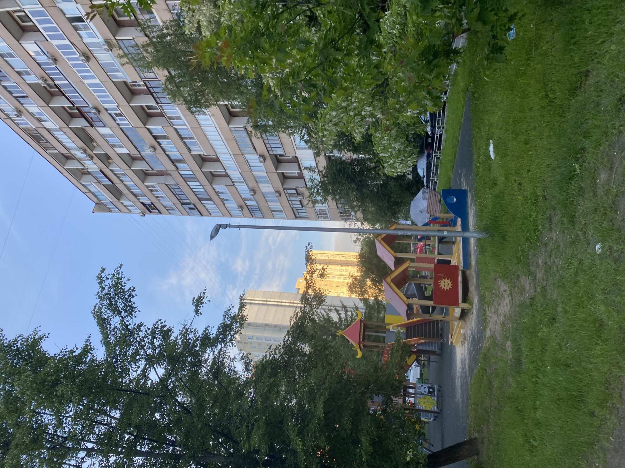 This image depicts an outdoor scene within a residential area in Khimki, Russia. The composition features a children's playground in the mid-ground on the right, surrounded by lush greenery and towering apartment buildings.
A tall, multi-story apartment building, characterized by numerous windows and balconies, dominates the left side of the frame. Its light-colored facade is partly obscured by dense green trees. Further in the background, other residential buildings are visible, including a gray-toned block and another building with a yellowish hue, illuminated by direct sunlight, suggesting either late afternoon or early morning.
The children's playground, located in the mid-ground on the right, is a colorful wooden structure with red and yellow accents, featuring slides and climbing elements. A prominent detail on the playground is a red panel adorned with a yellow, stylized sun emblem. A blue bench or seating area is also visible near the play structure. Adjacent to the playground is a paved pathway, bordered by a strip of neatly maintained green grass on the far right.
Numerous trees with vibrant green leaves are scattered throughout the scene, providing shade and depth. A tall street lamp pole extends towards the clear blue sky, which shows a few scattered white clouds. No people are visible, indicating a quiet moment. The clear sky and specific lighting suggest pleasant, possibly sunny, weather. The scene conveys a calm atmosphere typical of an urban park or courtyard.
