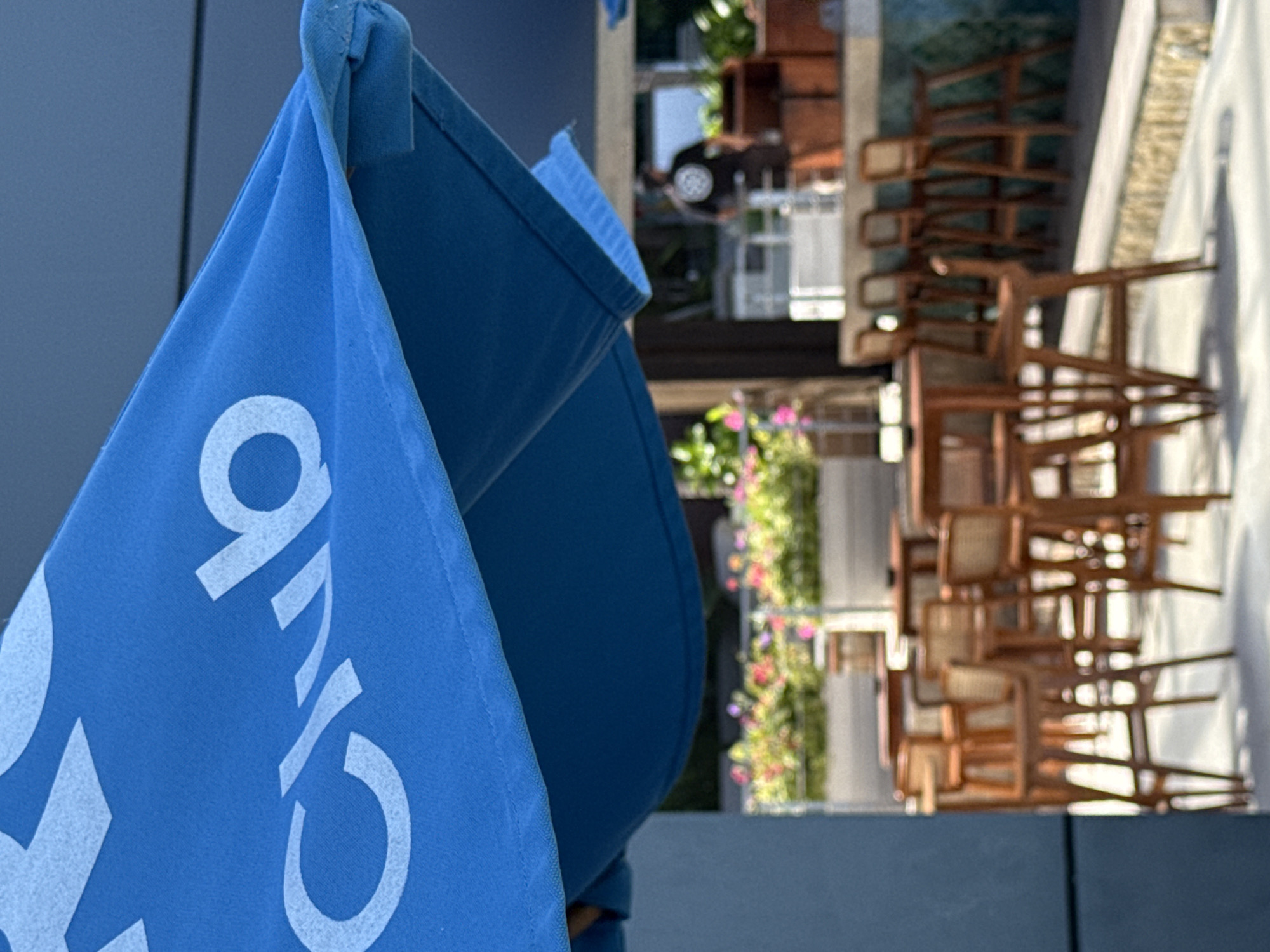 An outdoor seating area with a blue umbrella in the foreground displays white lettering that reads "Club". In the background, there are several wooden bar stools and tables, a bar, and a person wearing a black t-shirt and shorts. Flower boxes with pink and yellow flowers are also visible. The scene is illuminated by natural light, suggesting it is daytime. The setting appears to be a resort or restaurant in a tropical location.