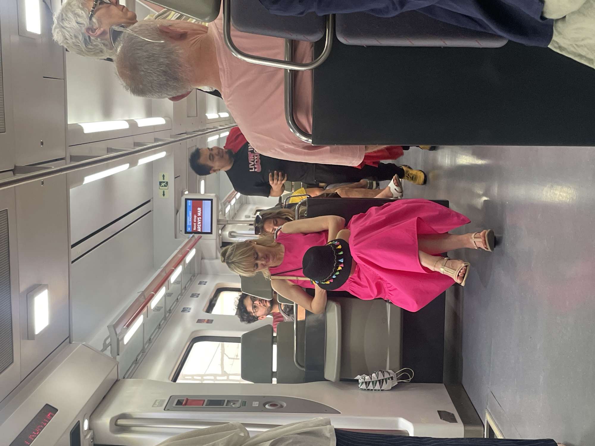 The image captures the interior of a modern train or subway car, populated with several passengers. The scene is illuminated by bright interior lighting, suggesting a well-lit environment.
In the prominent foreground, a woman wearing a striking hot pink dress and a black hat adorned with colorful tassels is seated, looking towards the right side of the frame. Her legs and feet, clad in light-colored sandals, are visible. She appears to be holding a thin, light-colored object in her hand.
Behind her, other passengers are visible. A man in a black t-shirt with white lettering (partially obscured, but appears to say "LIVING IN M...") is standing, looking right. Further back, another man with gray hair and a pink shirt is partially visible. An older woman with short, white curly hair and glasses is seated near the windows, facing forward. Another person with dark hair is also visible, seated further back.
The setting is a contemporary train car, characterized by clean white walls, a gray floor, and efficient strip lighting running along the ceiling and above the windows. Gray individual seats are arranged throughout the car. Horizontal handrails are mounted above the window line.
A digital display screen on the wall clearly indicates "NEXT STOP:" followed by "ARENYS MAR". Another screen shows "23 RAGAFFET". An illuminated green exit sign, featuring a white arrow pointing right and a walking figure, is also visible on the wall.
The overall activity is people traveling by train, with most passengers appearing relaxed or in transit. The environment is orderly and clean. The location context indicates this is likely a train in or around Sant Andreu De Llavaneres, Spain, with Arenys de Mar being a local destination.