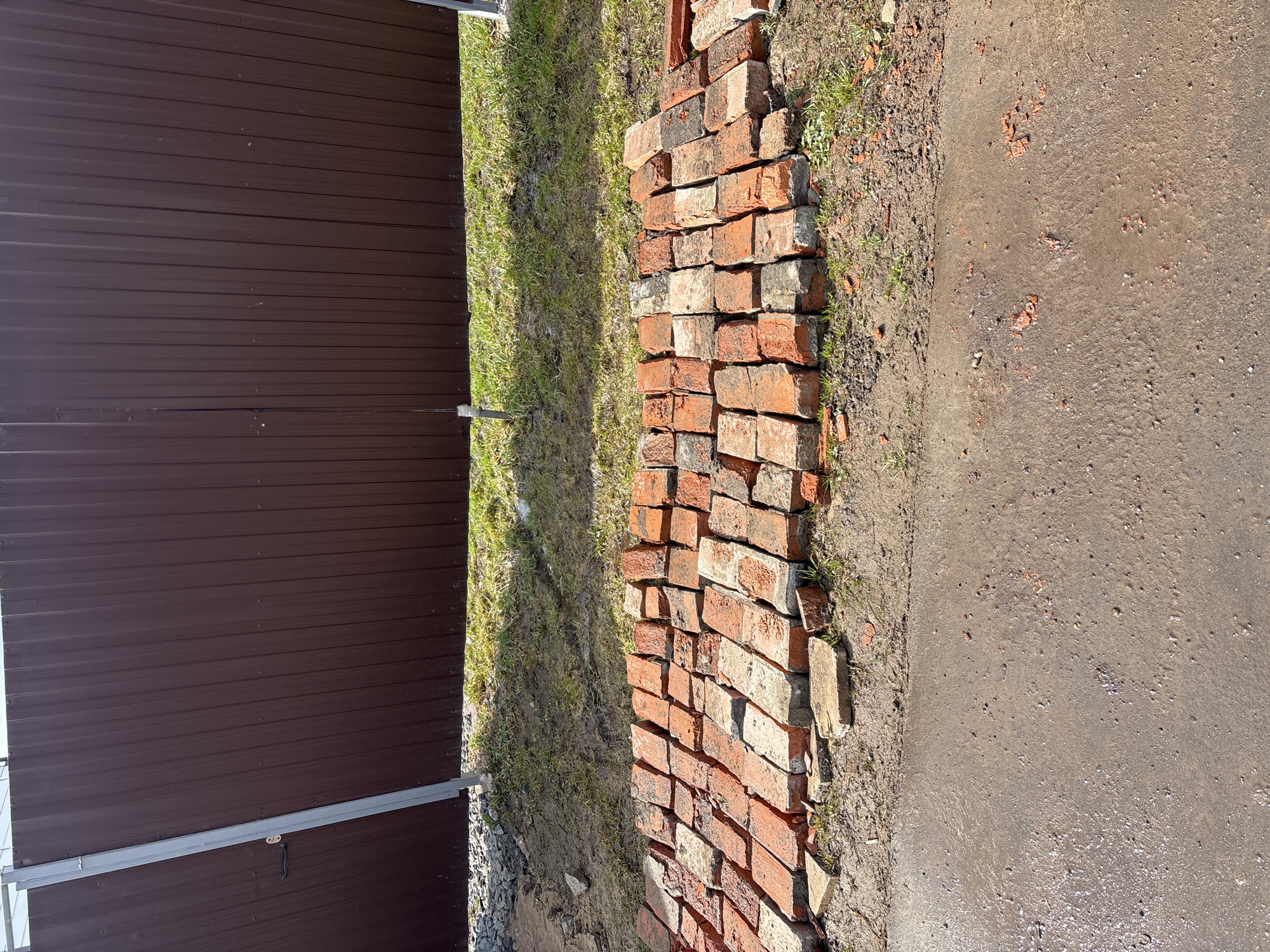 The image shows a pile of red bricks stacked unevenly on a patch of grass next to a brown metal fence. The grass is sparse and mixed with dirt. To the right of the bricks is a grey paved area. The image was taken during the day, with sunlight visible. The location is Раздолье, Russia.
