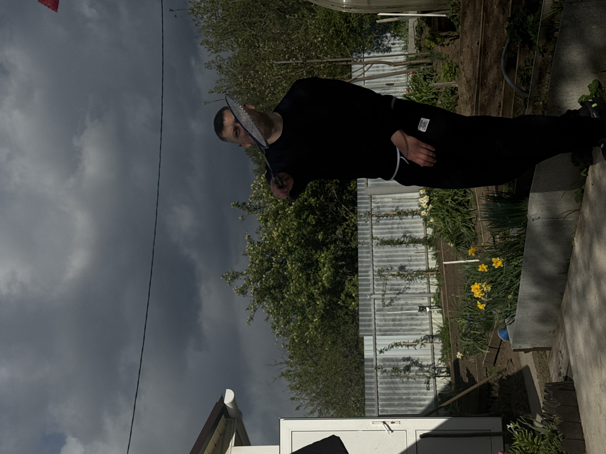 A person is playing badminton in a backyard in Chaikovsky, Russia. The person is dressed in black and is holding a badminton racket. There is a garden with yellow flowers and a fence in the background. The sky is cloudy.