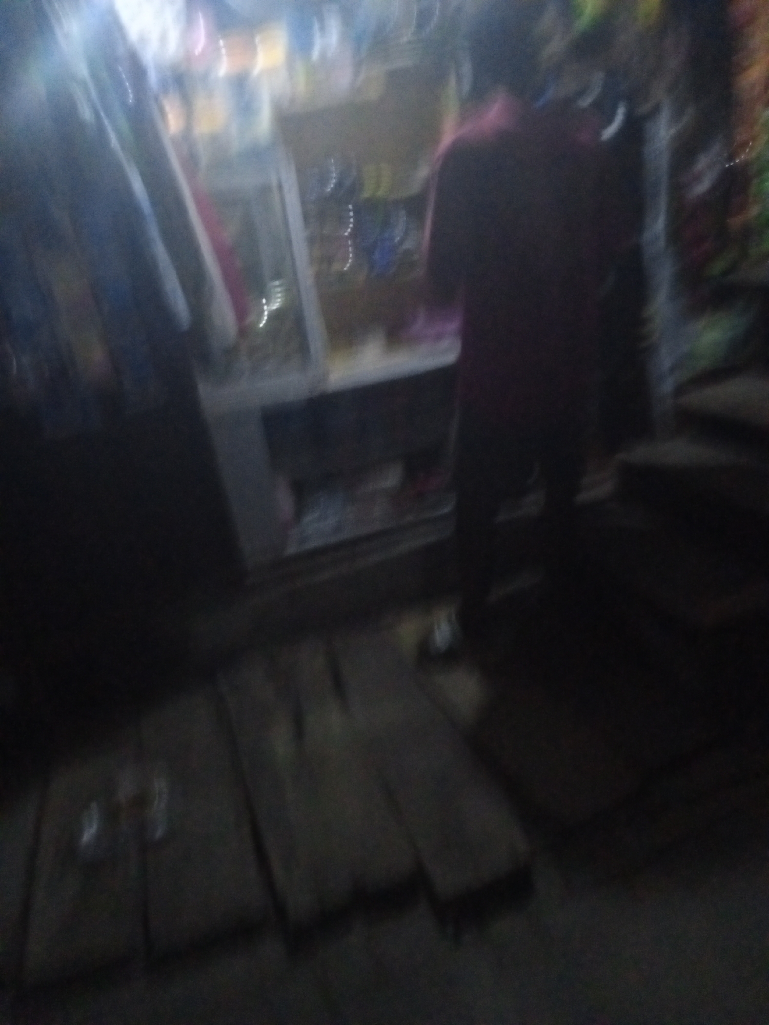The image shows the interior of a small shop in Ado, Nigeria. A person wearing a reddish-brown shirt stands near a counter. The shop appears to sell small items, with shelves displaying various goods. The floor is uneven and made of dark wooden planks. The lighting is dim, making it difficult to discern specific details.