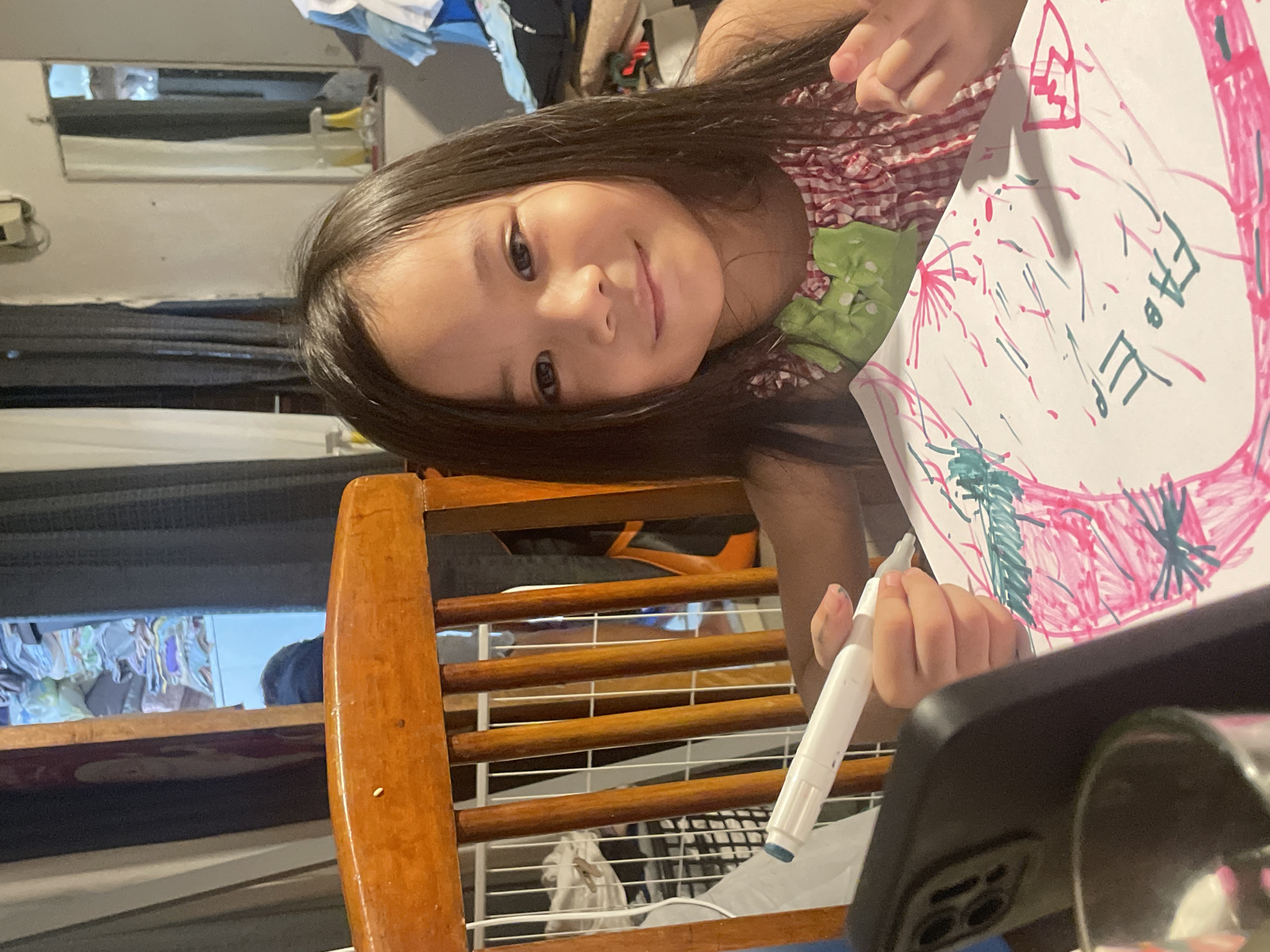 A young girl with long dark hair is sitting at a table, drawing on a piece of paper with a marker. She is wearing a red and white checkered dress with a green bow. In front of her is a smartphone. Behind her, a wooden chair is visible, and further in the background, there are curtains, a mirror, and shelves filled with clothes. The scene appears to be indoors, likely in a residential setting in Davao City, Philippines, given the context provided.