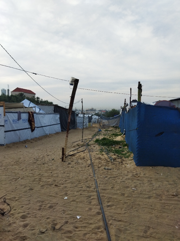 The photo depicts a refugee camp with makeshift shelters constructed from tarpaulins and various materials. The ground is sandy, with electrical wires running across the terrain. Wooden poles support the shelters and carry overhead lines. Some vegetation is seen between the shelters. A town or city is faintly visible in the background, suggesting the camp's proximity to a more developed area. The sky is overcast, giving the scene a somber tone.