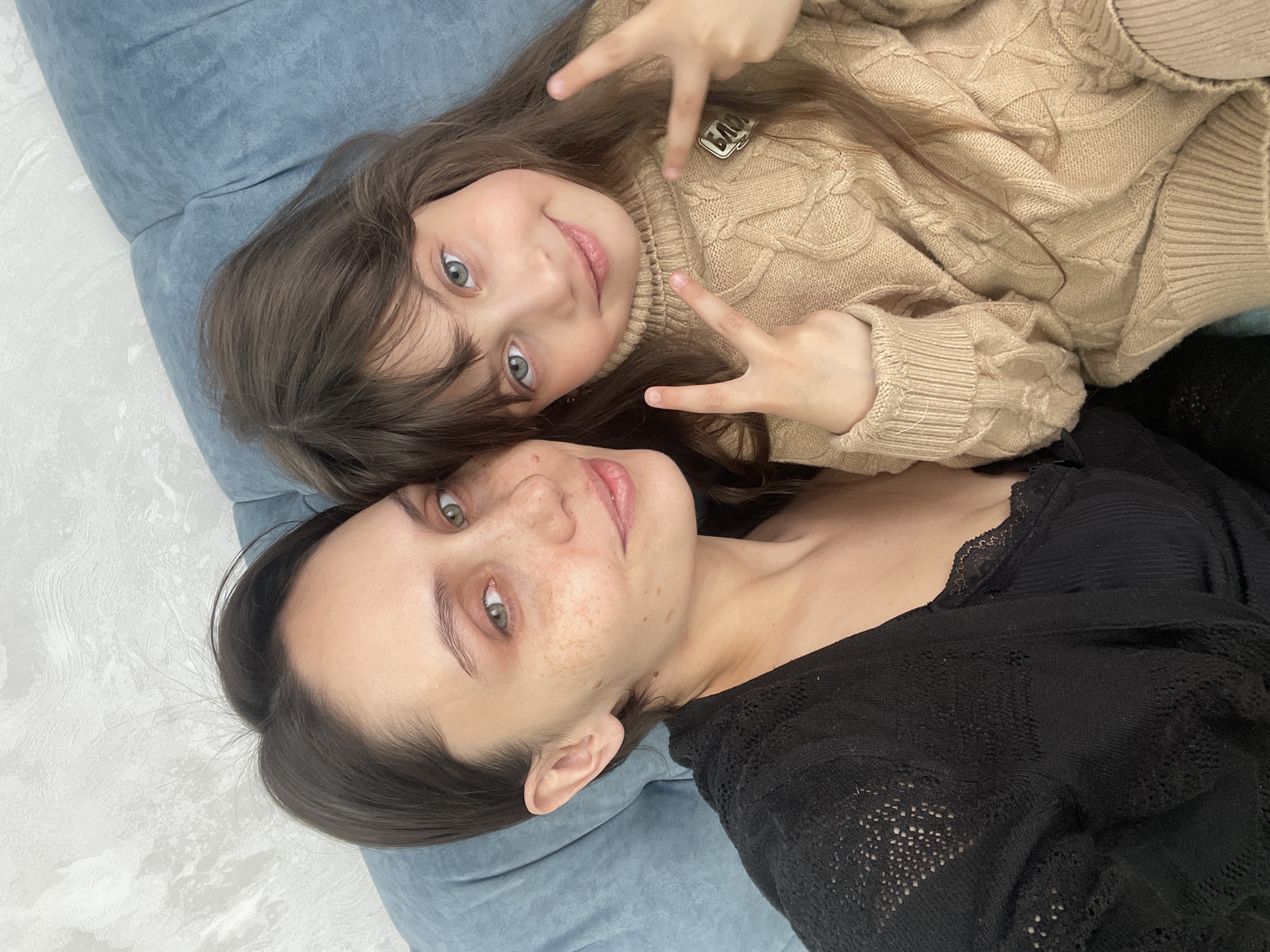 A woman and a child are lying on a blue surface. The child is wearing a light brown cable-knit sweater and making a peace sign with both hands. The woman is wearing a black knit top. The location is not identifiable as Izhevsk, Russia.