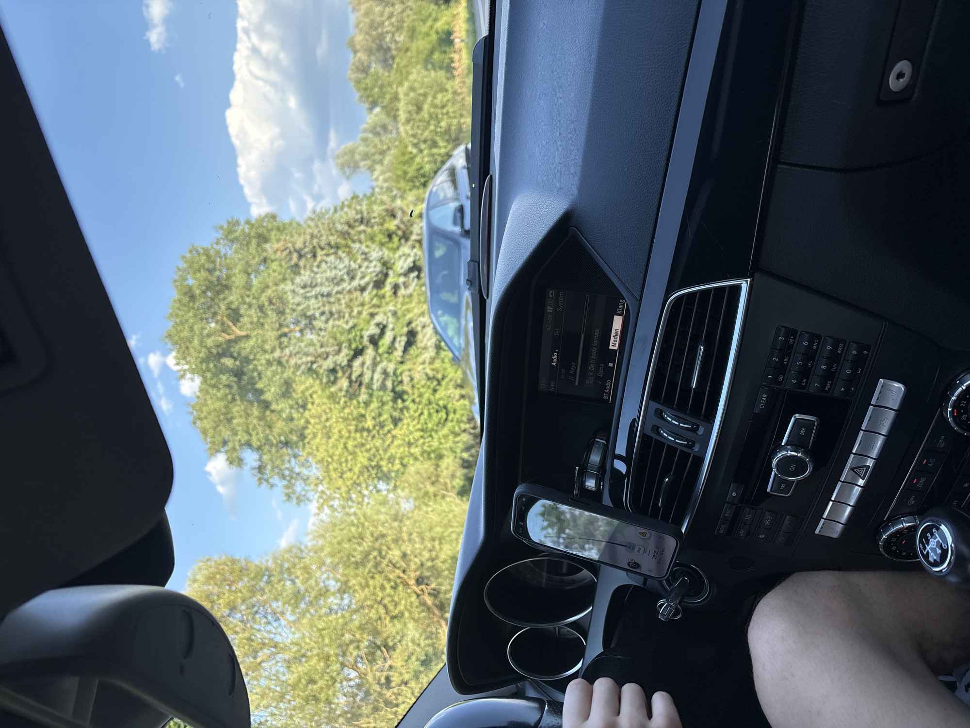 The image shows the interior of a car, looking out through the windshield. The car appears to be parked or driving slowly on a road bordered by trees and vegetation. A dark-colored car is visible in front of the vehicle. The dashboard of the car has a central display screen showing navigation and audio information, and a smartphone mounted on a holder. A person's arm and hand are visible resting on the steering wheel. The environment suggests a suburban or rural setting under a blue sky with scattered clouds. The location is identified as Iffezheim, Germany.