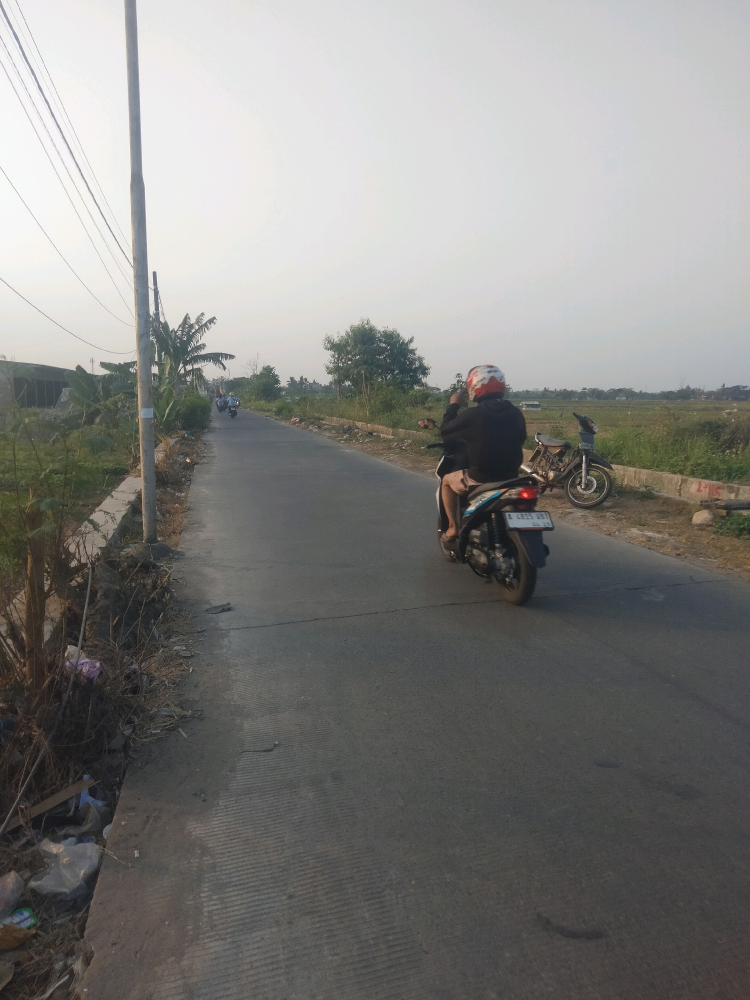 A person wearing a red and white helmet rides a scooter down a paved road in Suka Sari, Indonesia. The rider is wearing a black hooded jacket and light-colored shorts. The scooter is blue and black and has a license plate that reads "A 4875 UBT". In the background, another motorcycle is parked on the side of the road. The road is flanked by dry grass, a concrete barrier, and some vegetation. Power lines stretch across the sky, and a utility pole stands to the left of the road. The sky is overcast, and the overall lighting suggests it is late afternoon or early morning. There is some trash visible along the side of the road.