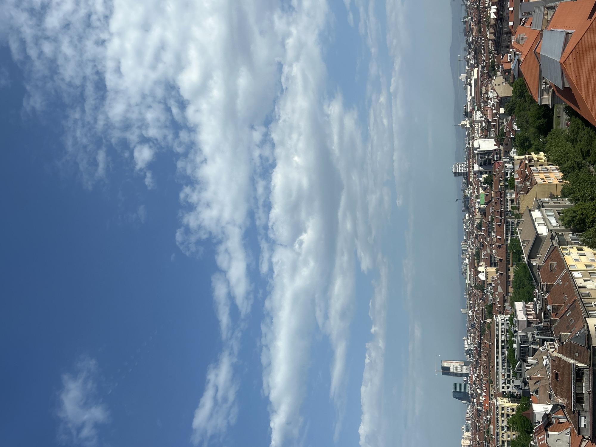 The image shows a high-angle view of Sofia, Bulgaria, on a sunny day with scattered clouds. The cityscape is characterized by buildings with red-tiled roofs, interspersed with trees and occasional taller modern buildings. The sky is predominantly blue with streaks of white clouds. In the distance, there is a hazy view.