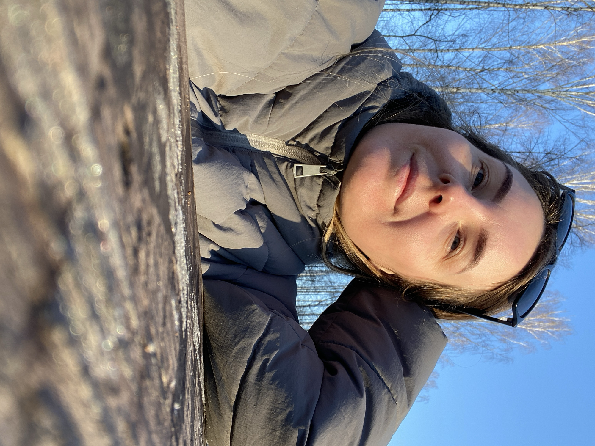 The image captures an adult female in a close-up, low-angle portrait, situated outdoors in the city of Noviy, Russia. The subject, with fair skin and light brown hair, is wearing a grey, possibly olive-tinted, puffer jacket with a partially visible metal zipper. Black sunglasses are positioned on top of her head. She is looking directly towards the camera with a slight, closed-mouth smile, and appears to be resting her upper body on a surface, with her right arm (viewer's left) bent and supporting her head. The immediate foreground consists of a dark, textured wooden surface, out of focus, showing some lighter residue along its edge. The background reveals a clear, bright blue sky above a line of tall, slender, bare deciduous trees, suggesting a late autumn or winter environment. The scene is illuminated by direct sunlight.