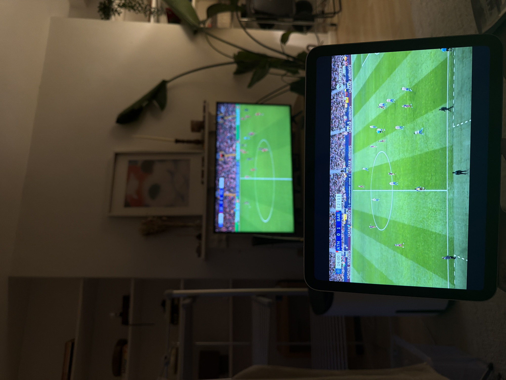 The image shows two screens displaying a football match. A larger screen in the background displays a broad view of the field, while a tablet-like device in the foreground shows a closer, more detailed view of the same game. The tablet shows the score as ATM 0-1 BAR at 14:00 minutes, referencing a Champions League match.
The background includes elements of a domestic setting, such as shelves, a picture frame, and a large plant, suggesting the scene is inside a home. Given the style of the interior and that the Champions League is a European competition, the location is likely somewhere in Europe, although this cannot be said with certainty from the image. It is not possible to determine if this is in the city of London, United Kingdom.
