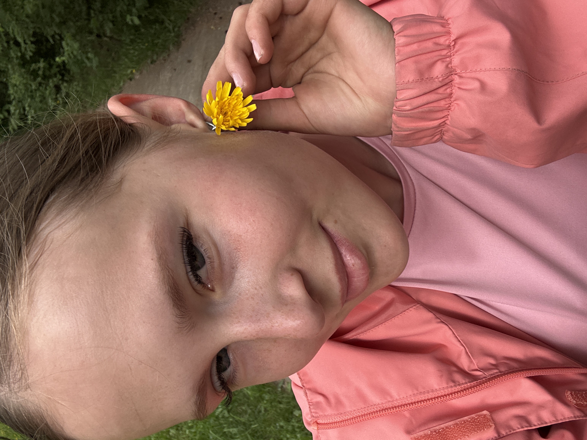 A young person with blonde hair pulled back is shown in a close-up selfie, holding a yellow dandelion near their right ear with their right hand. They are wearing a pink t-shirt and a coral-pink zippered jacket with Velcro closures. The background appears to be a path in a wooded or grassy area. There are no visible landmarks or indications of the specific location in Chaikovsky, Russia.