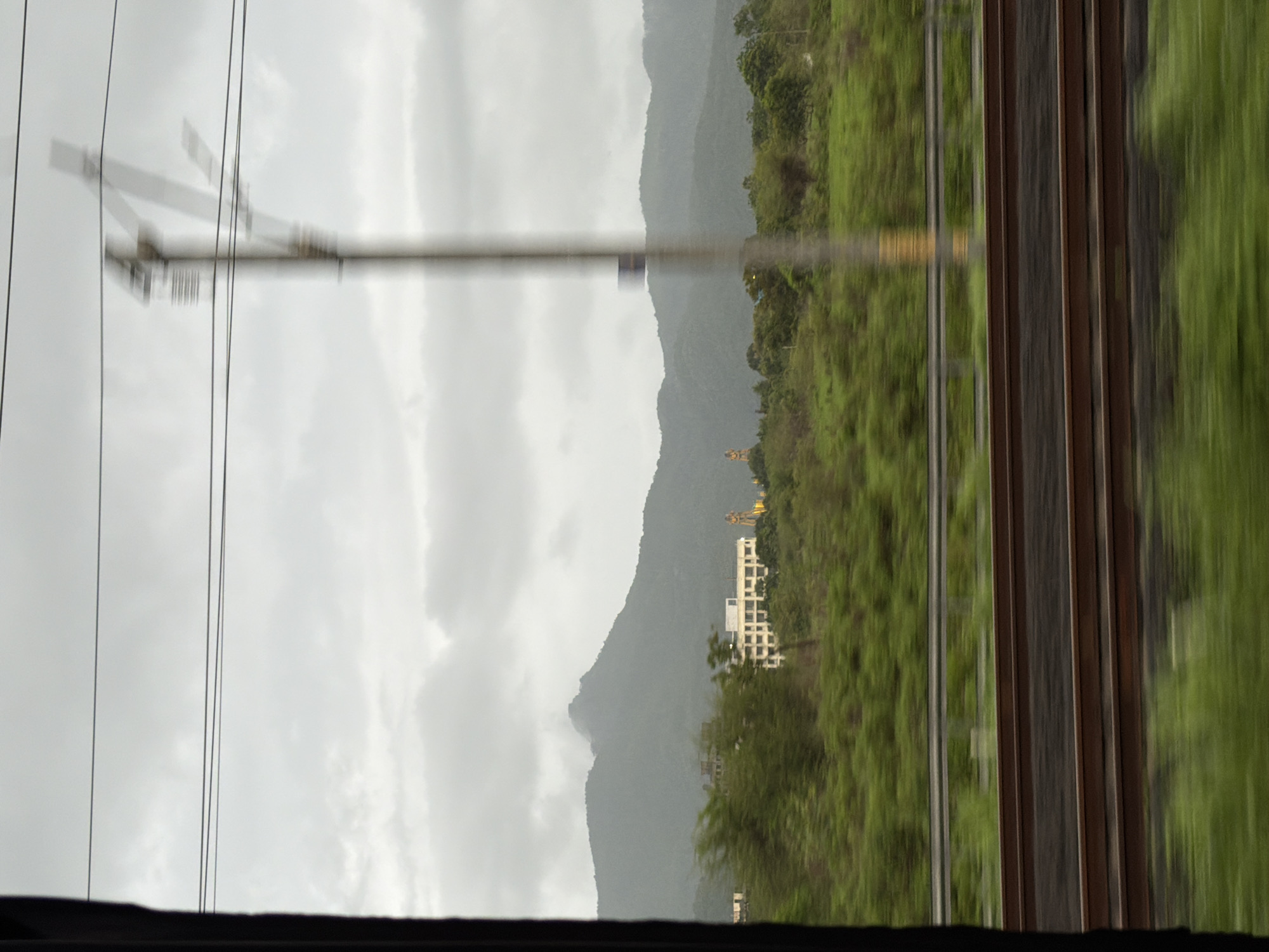The image depicts a view from a moving vehicle, likely a train, looking out at a landscape. In the foreground, a blurry section of train tracks runs horizontally across the frame. Beyond the tracks, a dense expanse of green foliage, possibly trees and bushes, stretches across the midground. A pale, almost white building with multiple windows is visible in the distance, situated amongst the greenery. Behind the building, a large, dark mountain range rises, its peaks shrouded in mist or clouds. The sky is overcast with a pale gray cloud cover. A blurry, vertical pole, possibly a utility pole, is present in the midground, partially obscuring the view of the landscape and sky. The overall impression is one of movement and a somewhat muted, possibly humid, atmosphere. This scene is consistent with the views often seen from trains traveling through the Palghar district of Maharashtra, India, which is characterized by its rolling hills, lush vegetation, and scattered settlements.