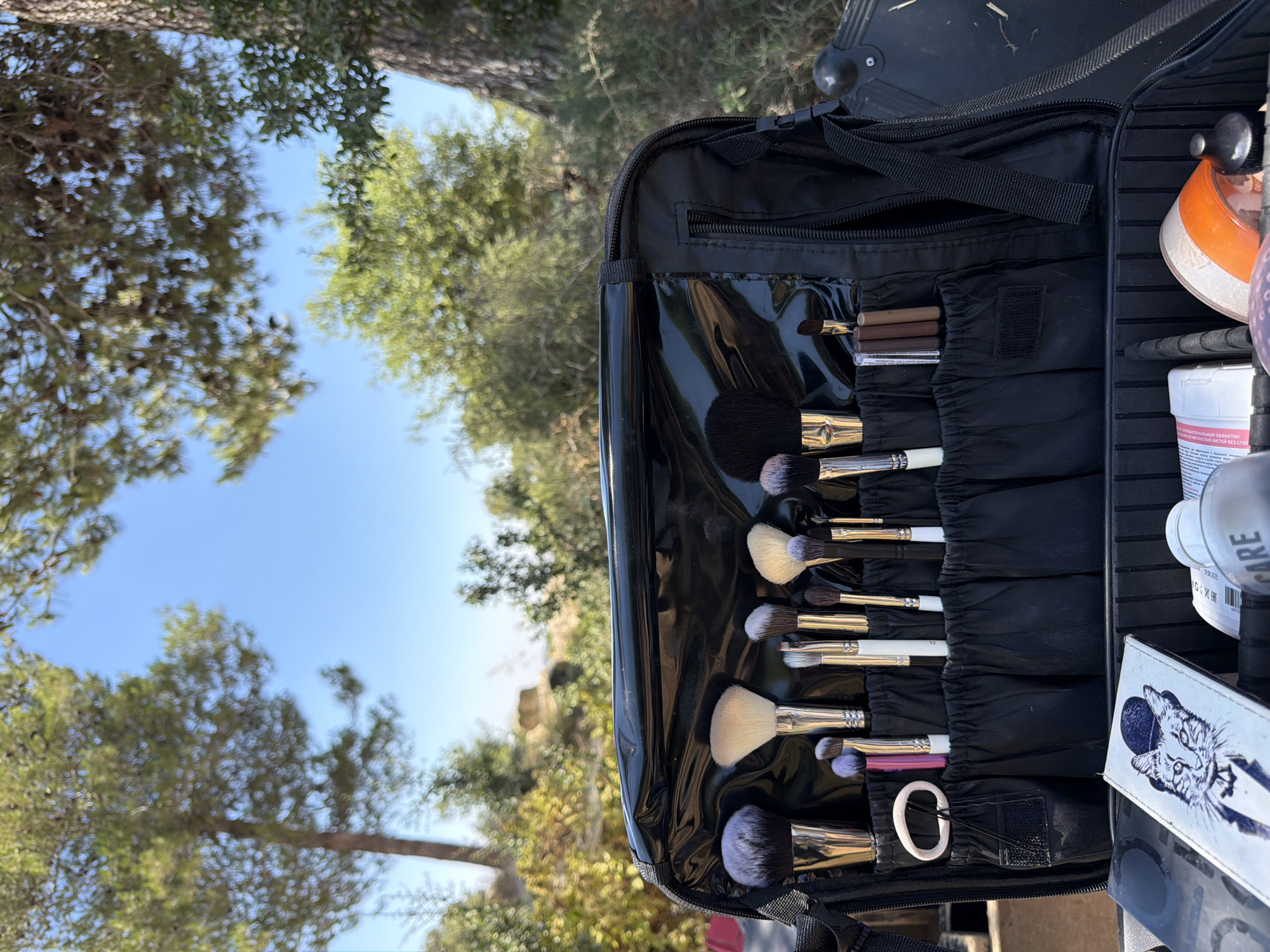 In Abu Gosh, Israel, a black makeup case is open, showcasing a variety of makeup brushes neatly organized in individual slots. The brushes have different sizes and colors, with some featuring black and silver handles and others white. Some brushes have purple tips. In the bottom compartment of the case, various makeup products are visible, including a white bottle with a pink label, a clear bottle, and a product with an orange lid. A white card with a drawing of a cat wearing a hat and bowtie is also placed in the bottom of the case. The case is positioned outdoors, with a background of green trees and a clear blue sky, suggesting a natural, possibly arid environment. The sun is shining brightly, casting shadows.