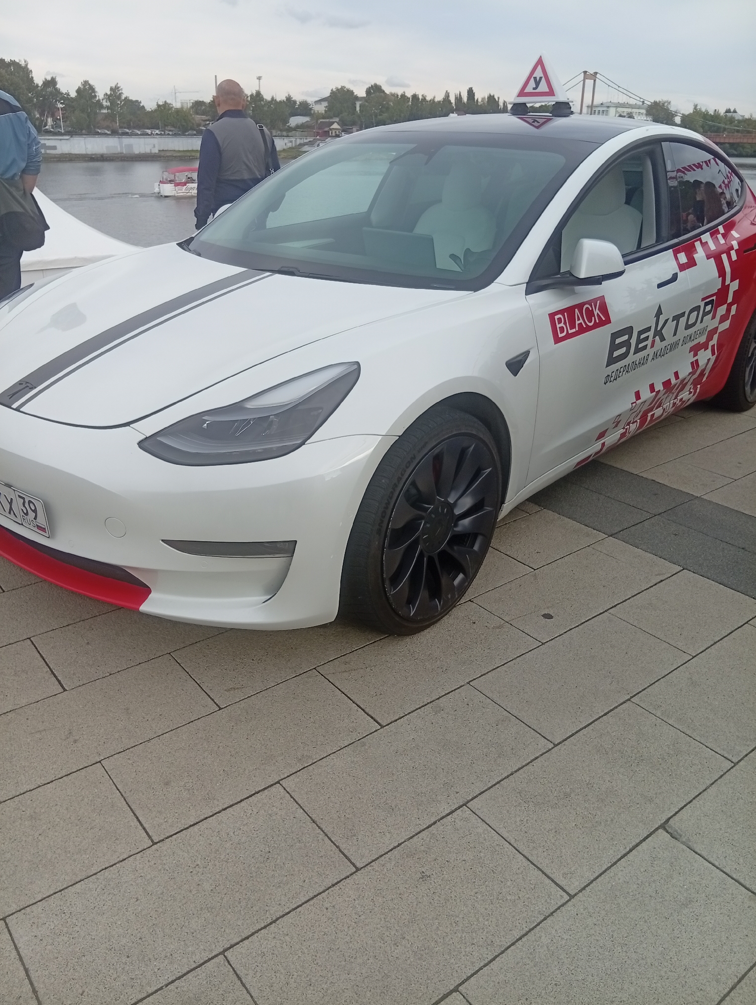 The image captures a white Tesla Model 3 parked on a paved walkway next to a body of water, possibly a river or canal. The car features racing stripes and a distinctive red and white graphic design on the rear quarter panel. A sign with a red border and a white "У" symbol, commonly used in Russia to denote learner drivers, is affixed to the car's roof. The car's license plate is visible and reads "KX 39 RUS". In the background, a small boat with people on board is visible on the water, and beyond that, trees and buildings line the shore. Two individuals are partially visible in the foreground, one wearing a blue jacket and the other a dark vest, standing near the car. The overall scene suggests an outdoor event or display, possibly related to driving education or a car show, in Penza, Russia, given the license plate format.