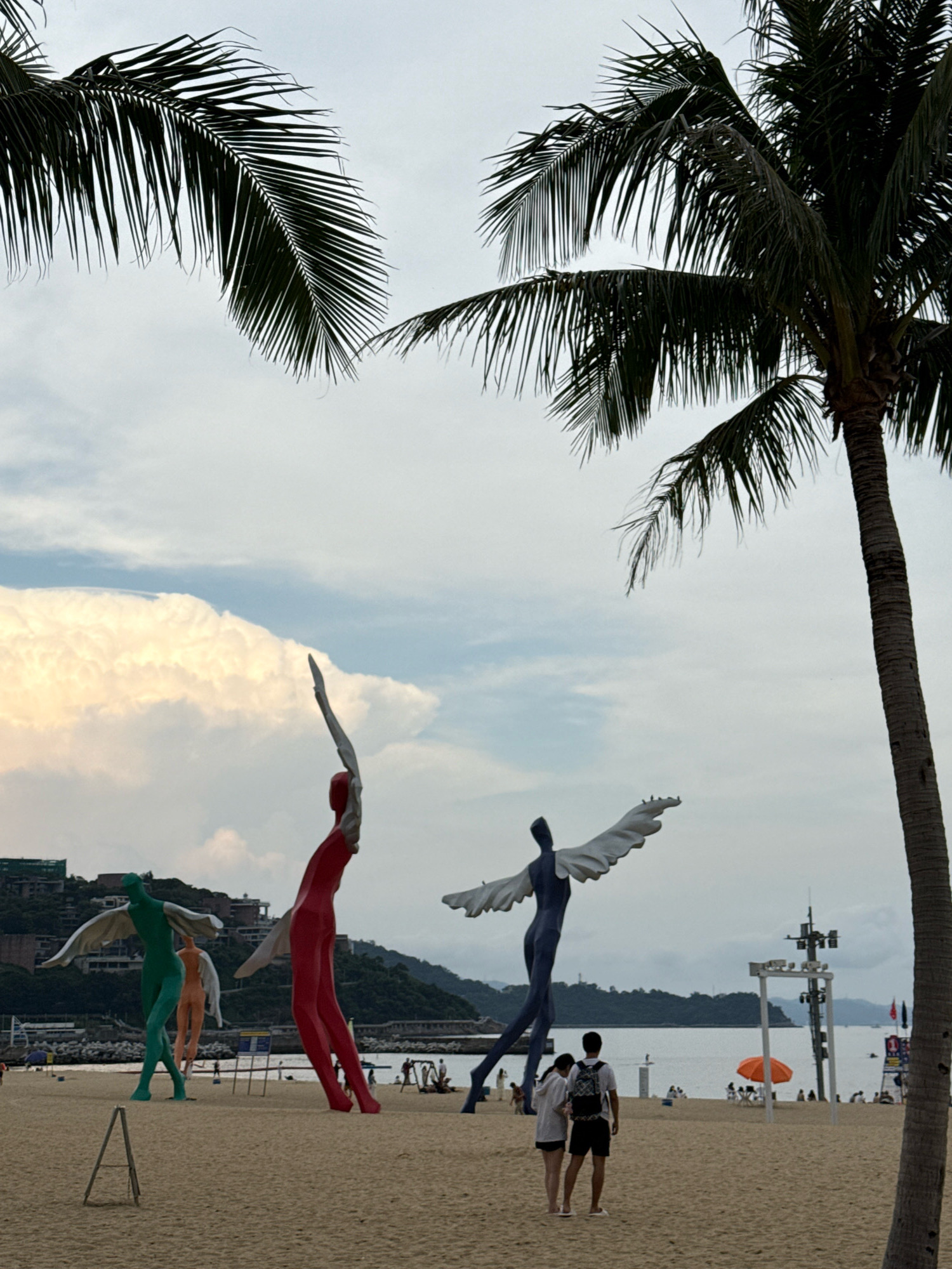 The image depicts a beach scene in Yantian District, China, characterized by its sandy shore and the presence of large, abstract human sculptures. Two palm trees frame the upper portion of the image, with their fronds extending into a cloudy sky. In the foreground, the sand is a light brown. Several towering sculptures are visible; one is colored red and is positioned to the right of center, with its arms raised. To its left, a green sculpture with outstretched wings stands, and further to the left, a partially visible orange-hued sculpture is present. In the mid-ground, a blue sculpture with white wings is depicted, and two individuals, a male and a female, stand facing away from the camera, in front of it. In the background, the ocean stretches towards a distant, hilly coastline dotted with buildings. A white structure with lights and an orange umbrella are also visible on the beach.