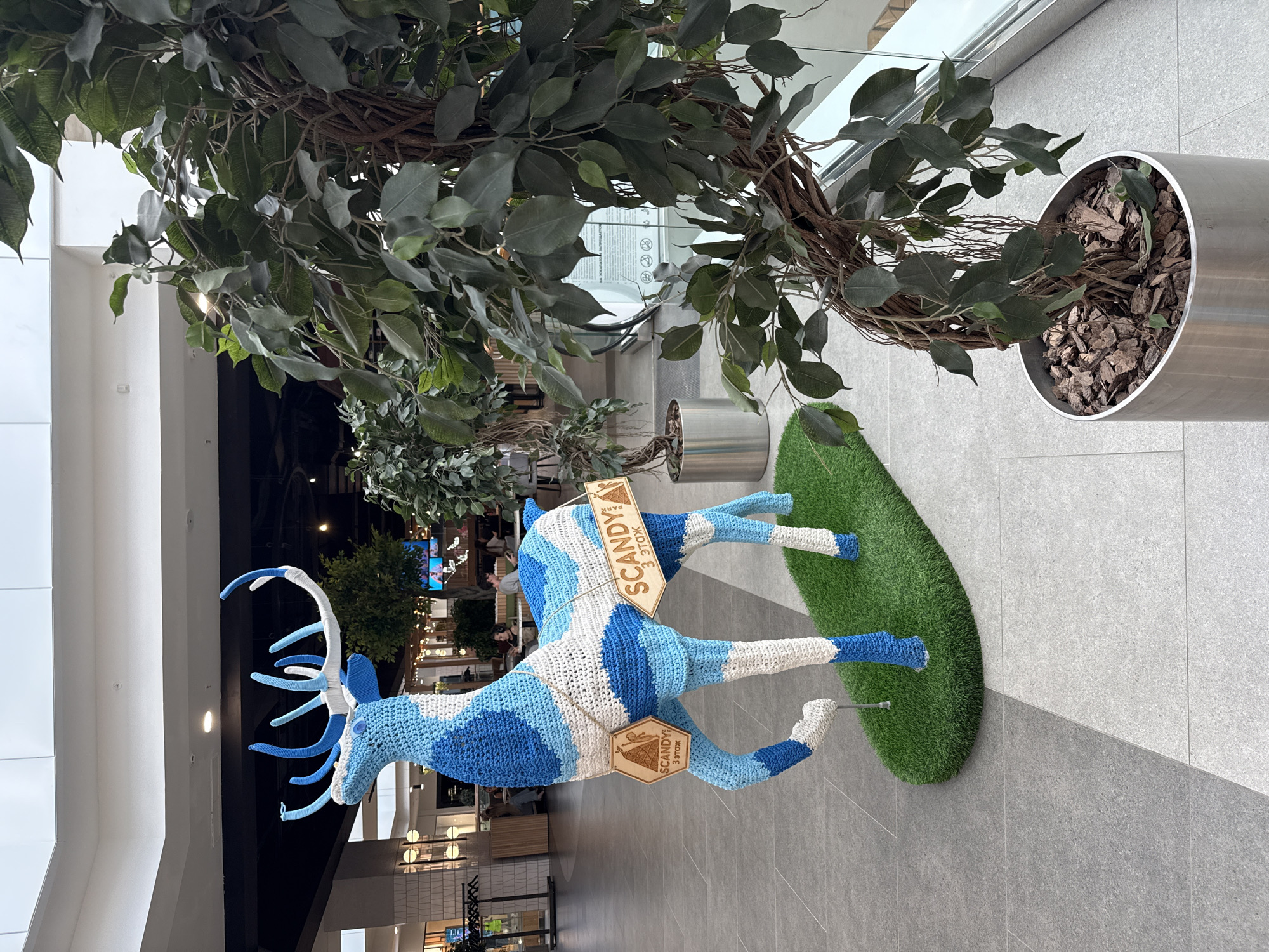 In Izhevsk, Russia, a knitted deer sculpture, adorned with blue and white patterns, stands on a patch of artificial grass in what appears to be a modern indoor shopping mall. The deer has prominent antlers and is decorated with two wooden signs that read "SCANDY PARK" and "3 этаж" (3rd floor). To the right of the deer, a large potted ficus tree with a twisted trunk and dark green leaves occupies a significant portion of the frame. In the background, the mall's architecture is visible, featuring light-colored floors, a high ceiling with a skylight, and shops with displays and people browsing. The overall scene depicts an interior commercial space with decorative elements.
