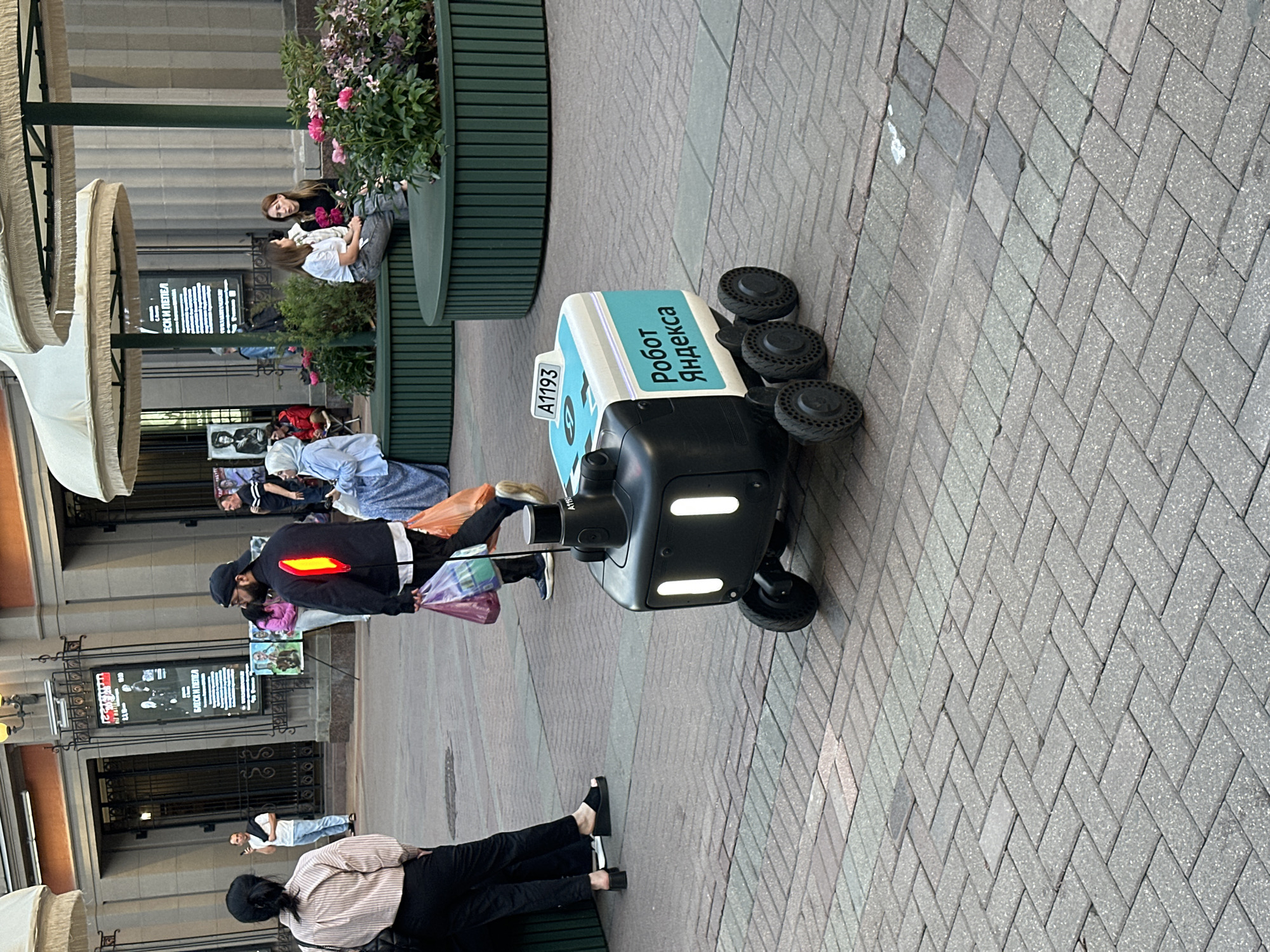 The image captures a vibrant urban street scene, likely in Moscow, Russia, given the presence of a Yandex delivery robot. The perspective is a slightly high-angle shot looking down a pedestrian thoroughfare.
The central subject is a Yandex delivery robot, light blue and black, with six wheels, situated on a paved sidewalk. The robot prominently displays "РОБОТ ЯНДЕКСА" (ROBOT YANDEX) in Cyrillic script and the identifier "A1193". Its front features two horizontal light strips.
Several people are visible. Near the robot, a person in a dark jacket with a bright red reflective strip on the back is seen, possibly interacting with the robot or passing by, holding plastic bags. Further back, two women are seated on a dark green planter-bench, engaged in conversation amidst lush greenery and pink flowers. Other individuals are scattered throughout the background, walking or standing near storefronts and awnings. One person is seated in the foreground, partially visible.
The setting is a pedestrian-friendly area with a light gray herringbone pattern paving. On either side, buildings with classical architectural elements, such as ornate window grilles and columns, line the street. Several commercial signs and posters are visible on the buildings, including one with partially legible Cyrillic text "ЕСКИ И ПЕПЕЛА". Light-colored outdoor umbrellas or awnings extend from the buildings.
The time of day appears to be late afternoon or early evening, with soft, ambient lighting. The atmosphere is casual, depicting everyday urban life with people going about their routines and modern delivery technology integrated into the city landscape.
