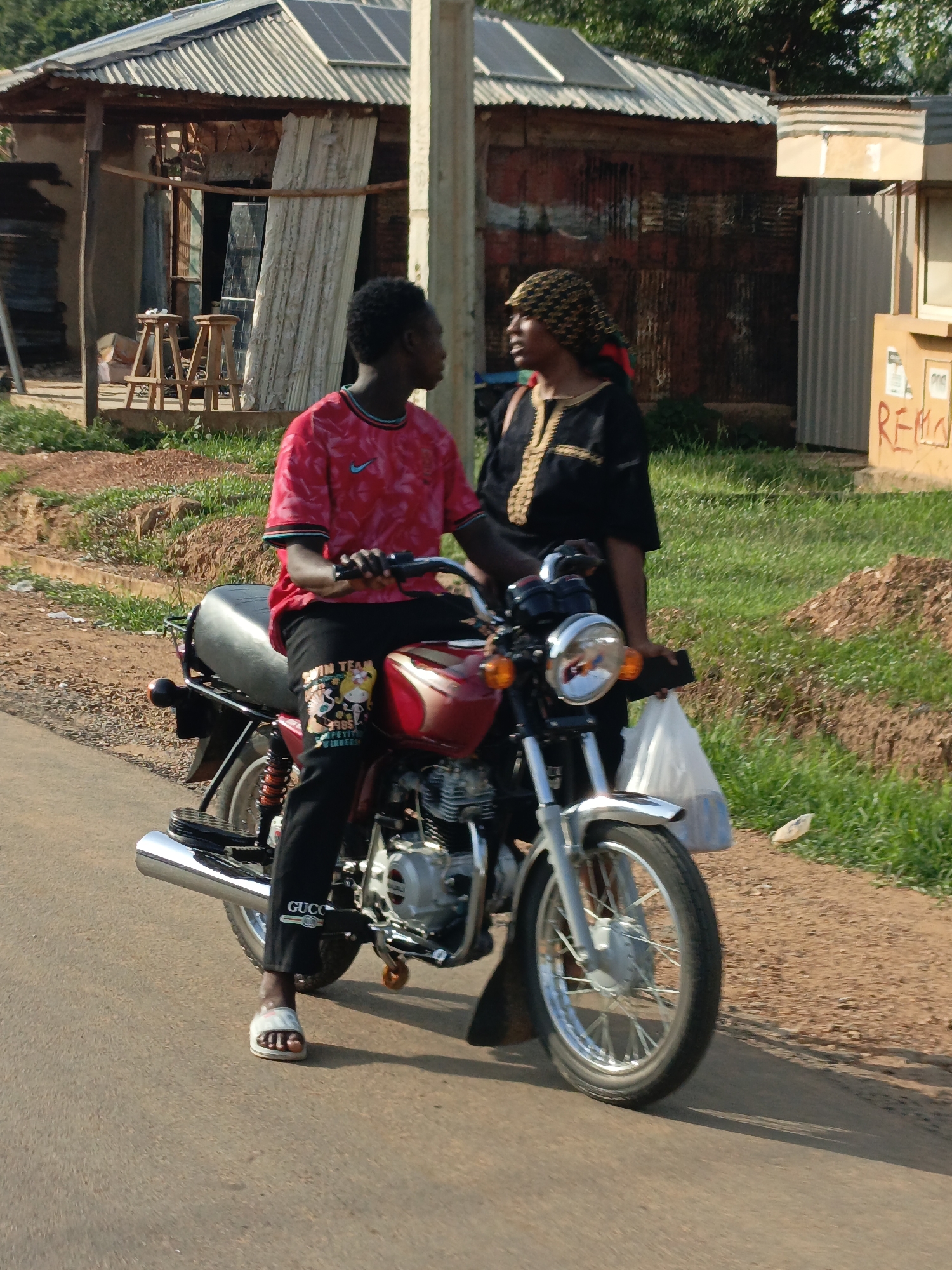 In Ube, Nigeria, a young man wearing a red shirt, black pants with cartoon images, and Gucci-branded slides sits on a red and silver motorcycle on a paved road. A woman with a patterned head covering, wearing a black dress with gold embroidery, stands beside him, holding a white plastic bag. They appear to be in conversation. In the background, there are buildings with corrugated metal roofs and a dirt shoulder covered with grass. A utility pole is visible near the buildings.