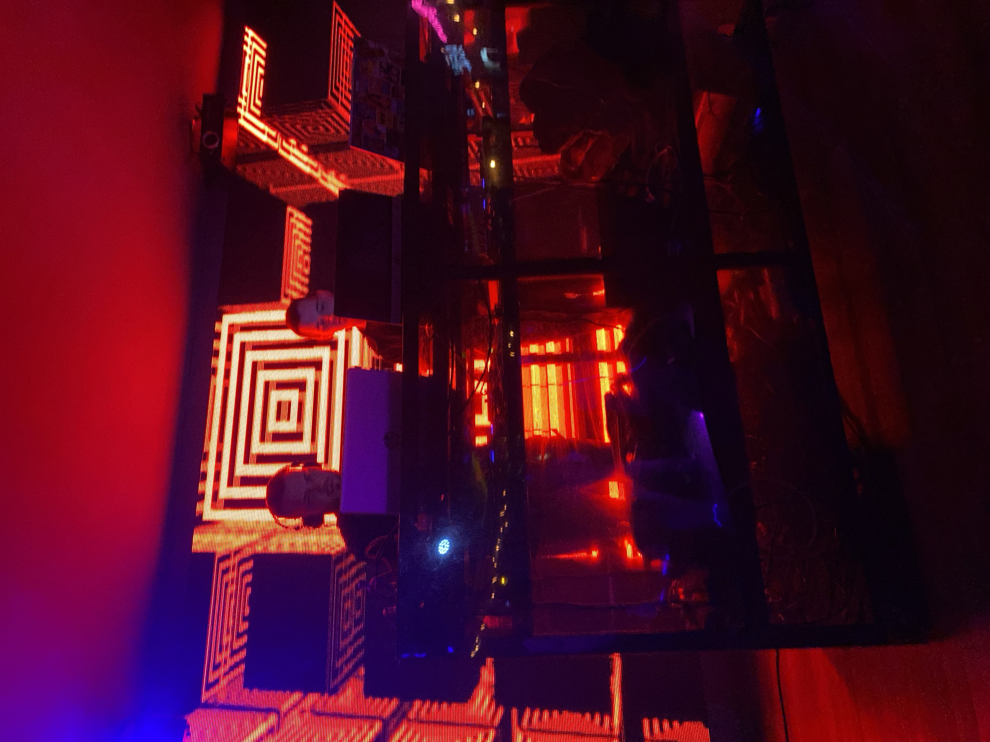 The image depicts a dark, indoor environment illuminated by strong, artificial red and blue lighting, suggesting a club, art installation, or futuristic setting. Geometric patterns of bright red and orange light, primarily concentric squares and horizontal lines, are prominently displayed on a dark surface on the left side of the frame, possibly a wall or screen.
A large, dark, reflective surface or glass panel occupies the central and right portions of the image, mirroring the illuminated patterns and other elements of the room, creating complex and distorted reflections. Within these reflections, faint outlines of what appear to be structures, wires, and additional light sources are visible.
Against the bright light patterns on the left, two human-like busts or heads are partially visible; one near the upper left and another towards the lower left. Their forms are indistinct, appearing as silhouettes or dimly lit figures. A small, circular, illuminated object, resembling a lens or a light, is mounted on a dark vertical surface near the upper light pattern. The overall scene is characterized by deep shadows, vibrant neon hues, and reflective surfaces, creating an intense, high-tech, and abstract atmosphere. There is no clear activity taking place, and no visible text is present. The artificial lighting suggests it is either night or an enclosed space without natural light.