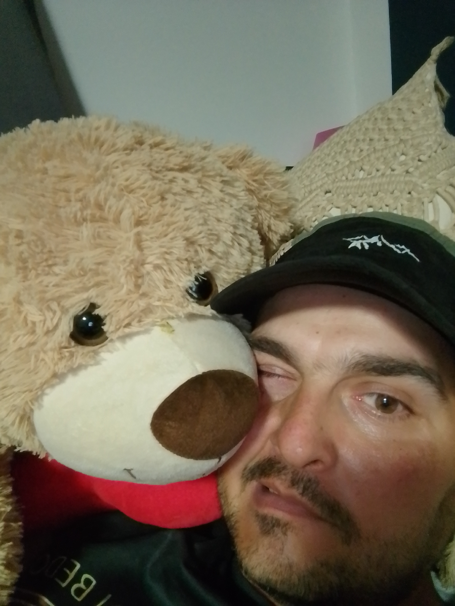 A close-up, selfie-style image shows a man and a large teddy bear. The man is wearing a black baseball cap with a white embroidered logo. His right eye is partially closed, and he has a mustache and goatee. The teddy bear, which is light brown and fluffy, is positioned next to the man's face, with its head resting near his left cheek. The background is out of focus but appears to be indoors with some textured fabric visible. The image was taken in Wedel, Germany.
