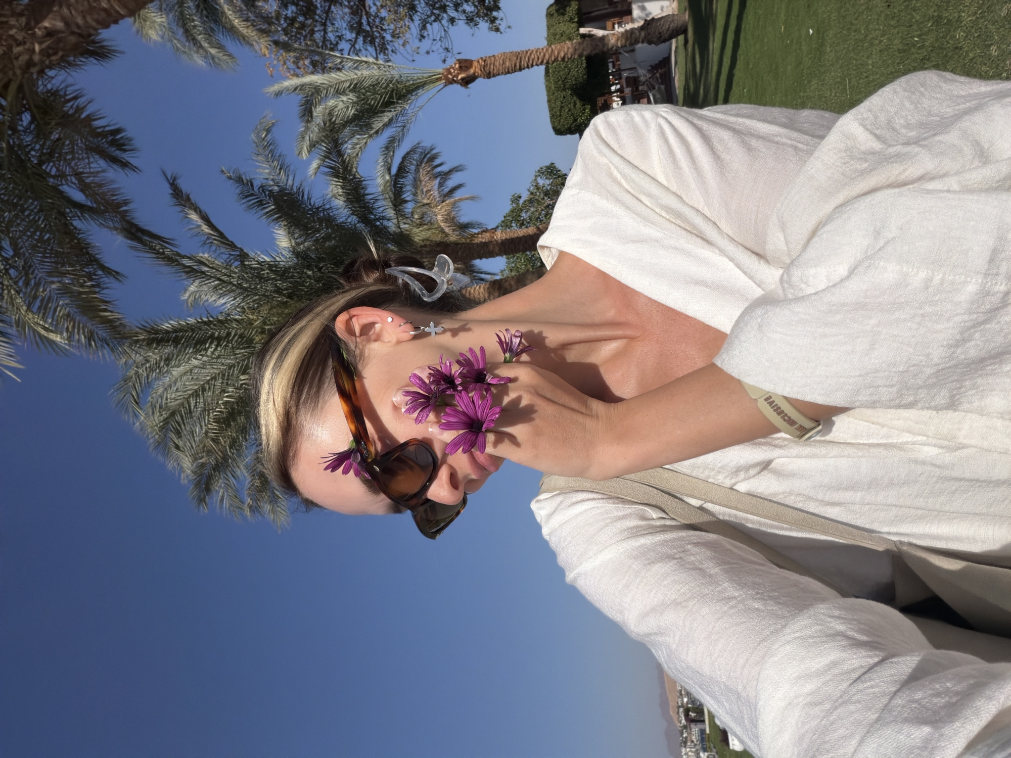 This bright, sunlit outdoor portrait captures a person, likely a woman, in profile, angled slightly downwards and to the left. She wears dark, tortoiseshell-framed sunglasses and holds several vibrant purple daisy-like flowers near her face, with some tucked into the sunglass frame. Her hair, featuring blonde highlights, is pulled back, revealing a clear, butterfly-shaped hair clip and a star-shaped dangle earring on her left ear. She is dressed in a loose, textured white top, and a light-colored strap, possibly from a bag, crosses her shoulder. On her right wrist, a pale wristband is visible with the text "ALL EXCLUSIVE".
The background features a clear, vivid blue sky dominating the upper portion of the frame, indicating bright daytime weather. Several palm trees are scattered throughout, with their fronds silhouetted against the sky. A manicured green lawn is visible in the mid-ground, and in the distant background, low-rise buildings and faint outlines of mountains suggest a resort-like environment. The image evokes a relaxed, holiday mood in a sunny, tropical setting.