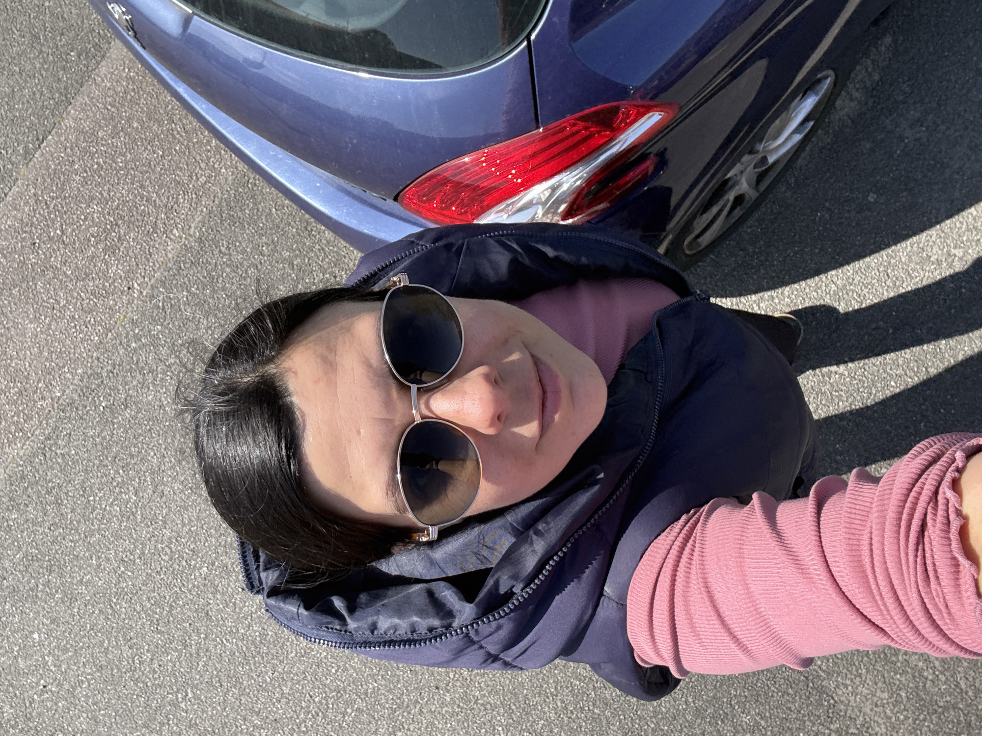 A woman wearing sunglasses and a pink shirt under a navy blue puffy jacket stands next to the rear of a dark blue car on a gray asphalt surface. The location cannot be determined to be in Great Yarmouth with the given information.