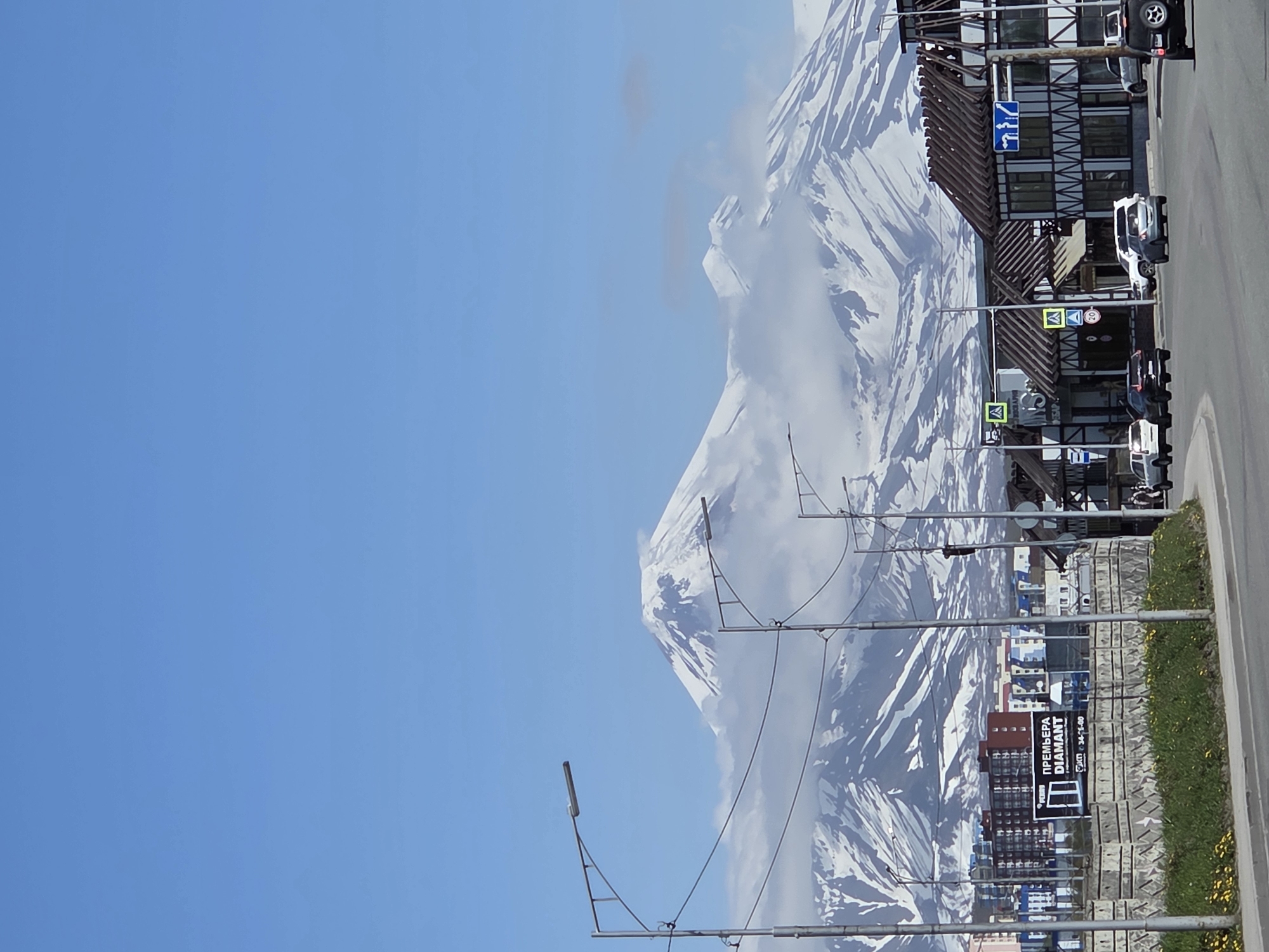 In an urban setting with a clear blue sky, a row of buildings lines a street. The buildings feature dark wood beams against white facades, with some displaying signage. A few cars are visible on the road, some parked and others in motion. Across the street, a concrete barrier is topped with grass and yellow flowers. In the background, a snow-capped mountain range dominates the skyline, partially obscured by clouds. The mountains appear rugged and majestic. Streetlights are present along the road, and power lines crisscross the scene. The overall impression is a blend of urban infrastructure and natural grandeur.