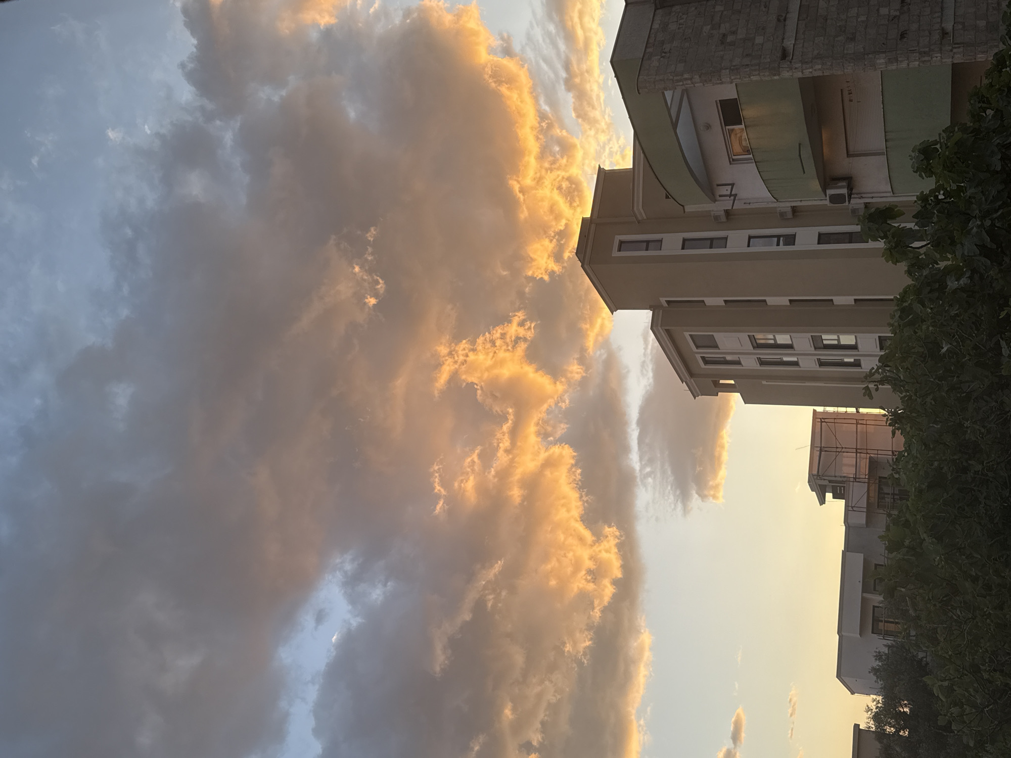 The image captures an urban or semi-urban landscape during either sunrise or sunset, dominated by a dramatic sky filled with large, textured clouds. The unseen sun casts a warm, golden and orange glow across significant portions of the clouds, creating striking highlights against areas of darker, grayish-blue cloud masses. A faint, slender crescent moon is visible in a small, clearer patch of bluish sky amidst the clouds in the upper left quadrant.
On the right side of the frame, several multi-story buildings are visible. The most prominent building, positioned centrally on the right, features a light, beige-toned facade with numerous windows. It has a distinctive architectural element on an upper floor: a curved, green canopy or awning. The top section of this building is clad in rough-hewn stone, and air conditioning units are affixed to its exterior. Further to the lower right, another building appears to be under construction or renovation, with scaffolding clearly visible around its upper floors. Lush, green foliage from trees or large bushes partially frames the right edge of the image, obscuring parts of the buildings.
The golden light illuminating the clouds is the most notable detail, strongly indicating either a sunrise or sunset, which, combined with the visible moon, creates a serene yet dynamic atmosphere. The architecture and visible development (scaffolding) suggest an active urban environment, consistent with the specified location of Bar, Montenegro. There are no people or visible text in the image.