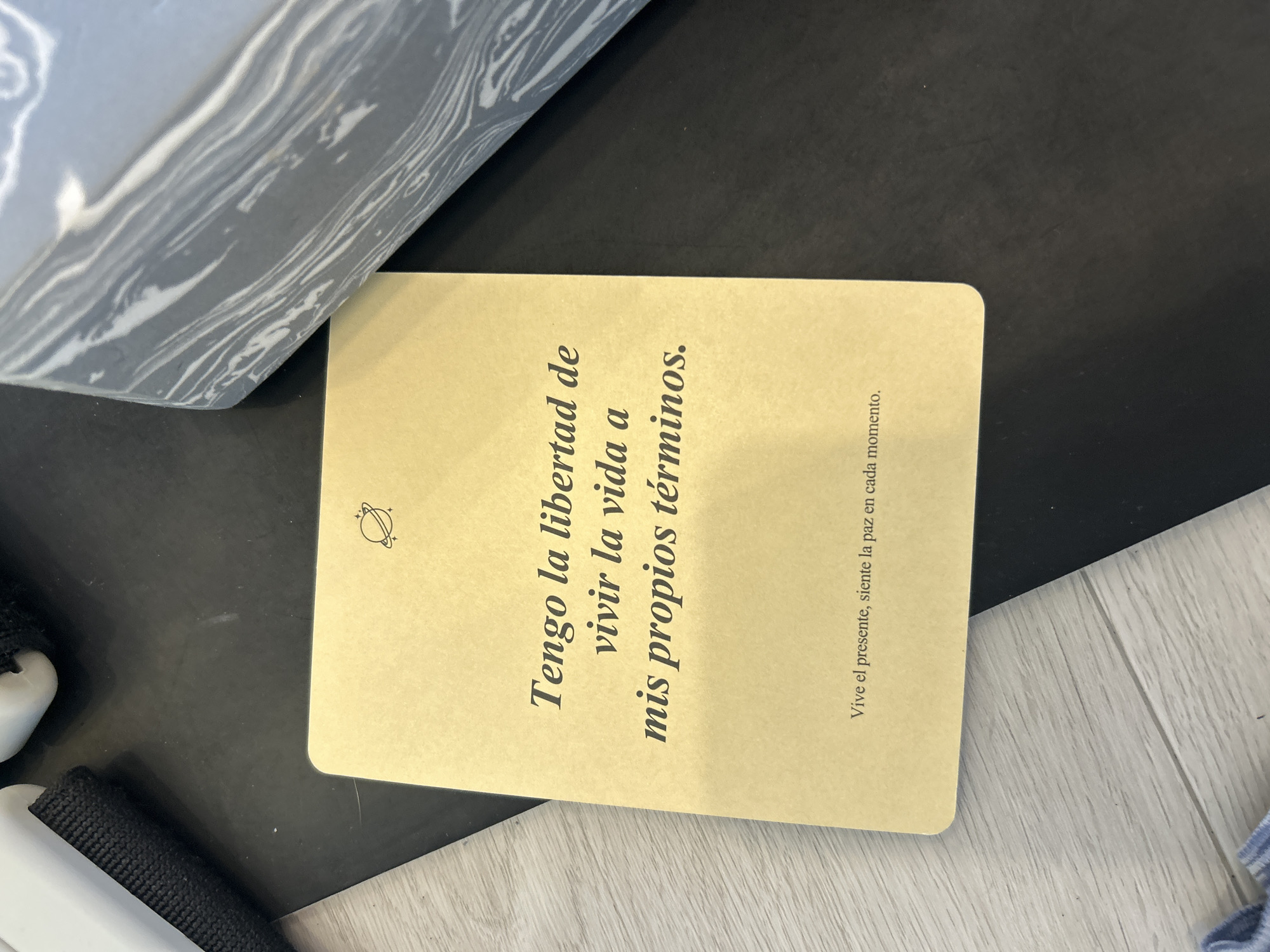 The image shows a rectangular card lying on a surface, partially obscured by an object with a gray marble-like pattern. The card is beige and has text in Spanish printed on it in a vertical orientation. The text reads: "Tengo la libertad de vivir la vida a mis propios términos." A smaller line of text reads: "Vive el presente, siente la paz en cada momento." There's a small icon above the main text that appears to be a stylized planet with stars. The background surface is a mix of black and light gray, with what appears to be a section of flooring visible in the lower right corner. To the left of the card, a black fabric strap with a white plastic component is visible. The location appears to be indoors but there is not any other information.