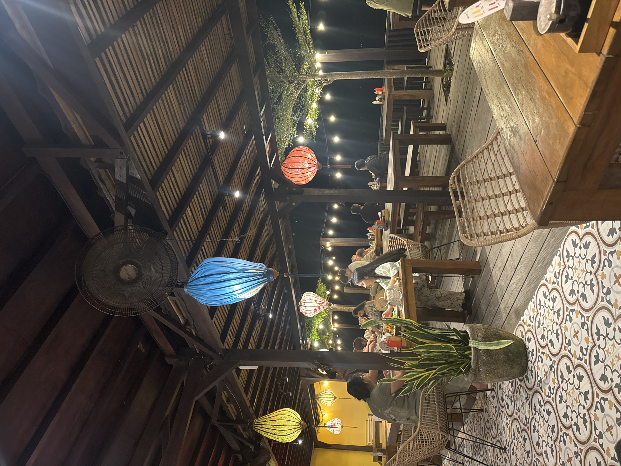 This inviting image captures an outdoor or semi-outdoor restaurant at night in Nusa Penida, Indonesia. The setting features a rustic wooden roof structure with exposed beams and slatted panels, creating an open and airy ambiance. Numerous string lights are strung across the ceiling and outdoor areas, complementing the warm glow emanating from several colorful lanterns in shades of blue, red, yellow, and white with patterns, hanging throughout the space.
Several people are visible, casually seated at wooden tables with chairs that feature woven rattan backs, engaged in dining and conversation. Their relaxed postures suggest a pleasant evening outing. A large, dark ceiling fan is prominently seen on the left, near a vibrant blue lantern. The floor transitions from wooden planks under the dining tables to decorative patterned tiles on the right side of the image, showcasing intricate motifs in brown, grey, and cream. A potted snake plant in a textured grey pot sits on these tiles, adding a touch of greenery. Beyond the covered area, more string lights are visible against the dark night sky, along with the silhouette of trees, reinforcing the outdoor setting. The overall mood is warm, cozy, and festive, indicative of an enjoyable evening atmosphere. No discernible text is clearly visible on any signs or menus.