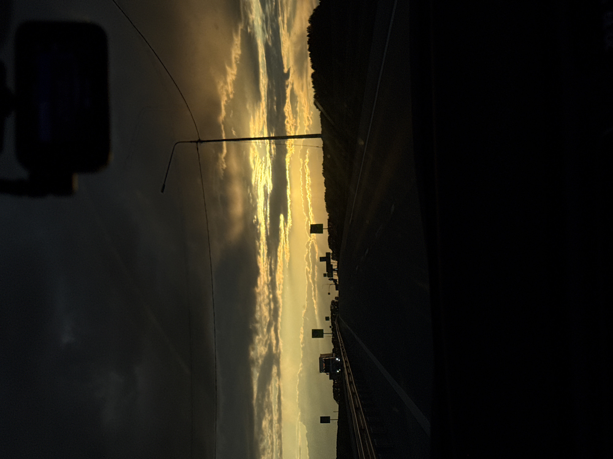 The image depicts a highway at sunset. The view is from inside a vehicle, likely a car, as suggested by the side mirror visible in the left foreground. The highway stretches into the distance, lined with trees on the right side. Streetlights are visible along the road. The sky is filled with clouds illuminated by the setting sun. The exact location is unknown, but the scene suggests a suburban or rural area near a highway.