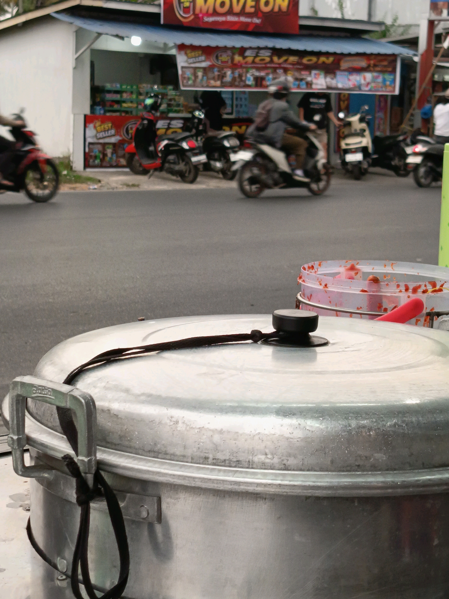 The image depicts a street scene in Banjarmasin, Indonesia. In the foreground, a large, silver metal pot with a handle and a black cord is prominent. Behind the pot, a clear plastic container with red sauce and a red object is visible. The background shows a street with several motorcycles parked and in motion. A small shop with signs such as "MOVE ON" and "BEST SELLER" is also visible. People are seen riding motorcycles, and one person is standing near the shop. The overall setting suggests a busy roadside area, possibly a market or a commercial street.