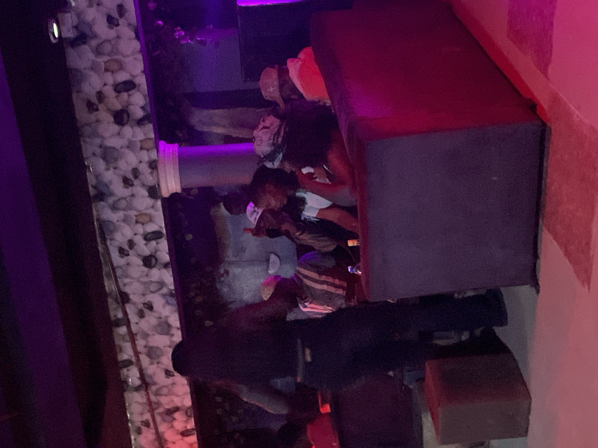The image depicts an indoor scene, likely a nightclub or lounge, characterized by dim lighting and purple and red ambient illumination. In the foreground, a large, dark-colored upholstered seating area or booth dominates the lower half of the frame. To the left of this seating, a light-colored square object, possibly a stool or a small table, is visible. The background features several individuals, some seated and some standing, amidst what appears to be a decorated wall with a pattern resembling stacked pebbles or stones. A tall, fluted column, illuminated in purple, suggests architectural elements. The overall atmosphere is social, with people appearing to be conversing and engaged. The location is identified as the city of Durimi, Nigeria.