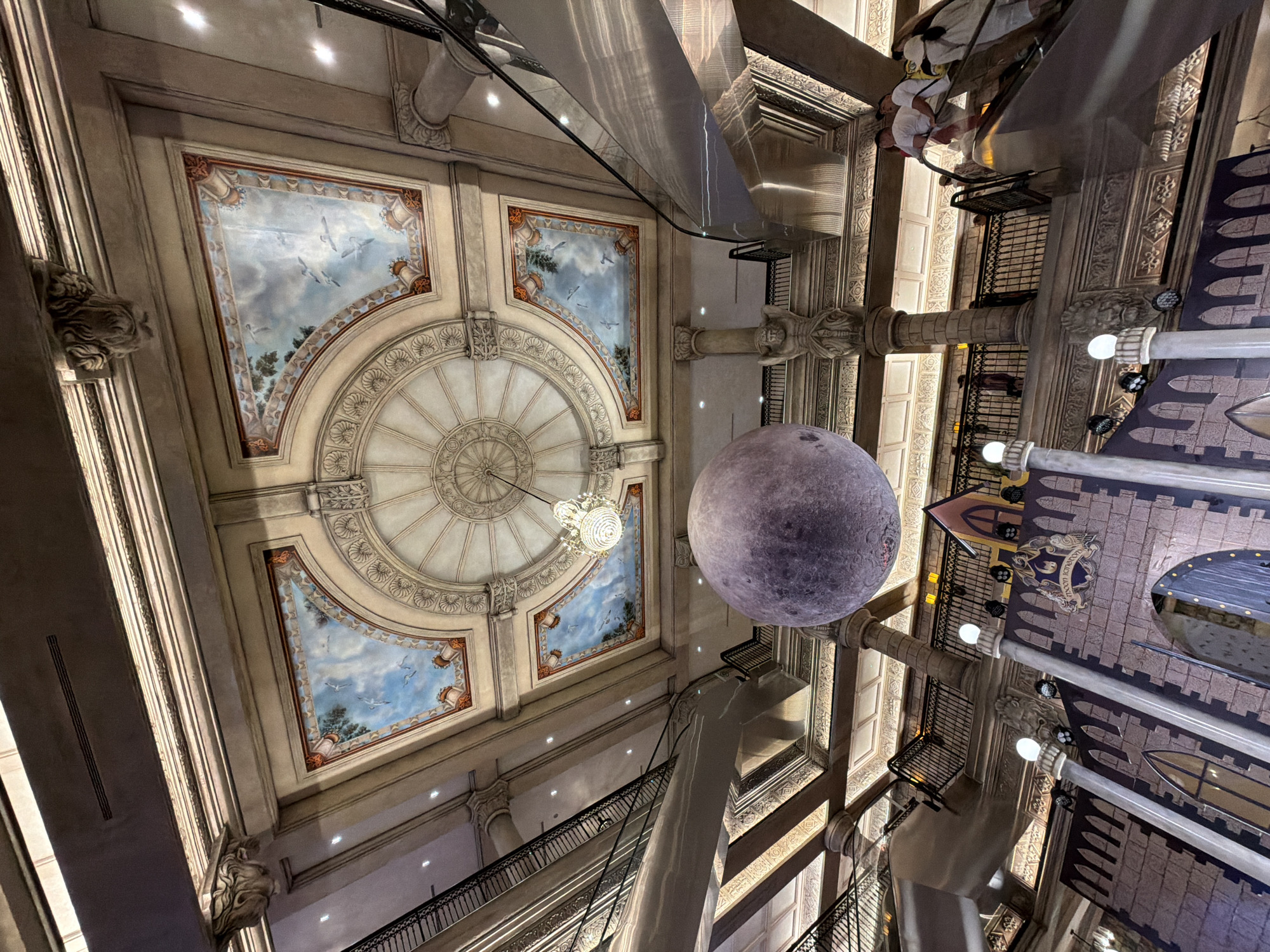 The image captures the grand interior of a multi-story building, likely a shopping mall or department store, in Da Nang, Vietnam. The scene is dominated by an impressive ornate ceiling adorned with painted murals of the sky and birds, framed by intricate moldings. A large, detailed sphere, resembling the moon, hangs suspended in the central atrium, illuminated by a crystal chandelier. Escalators and balconies with black railings line the upper levels, where several individuals, dressed in casual attire, are visible. The lower part of the image reveals a whimsical castle-like structure with a large stone facade and arched doorways, suggesting a themed display or event. The overall ambiance is one of opulence and imaginative decoration.