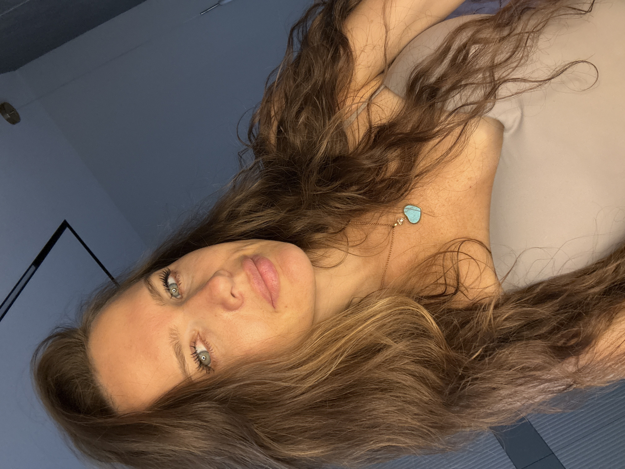 The image is a close-up, front-facing selfie of a woman with long, brown, wavy hair. She has light blue eyes, fair skin with some freckles, and full lips. She is wearing a light-colored top and a necklace with a heart-shaped turquoise pendant. The background is a plain, dark blue wall with some indistinct ceiling fixtures. The lighting appears to be artificial and comes from the side, casting shadows on her face. There is no indication of the specific location within the city of Khimki, Russia. The woman is looking directly at the camera with a neutral expression.
