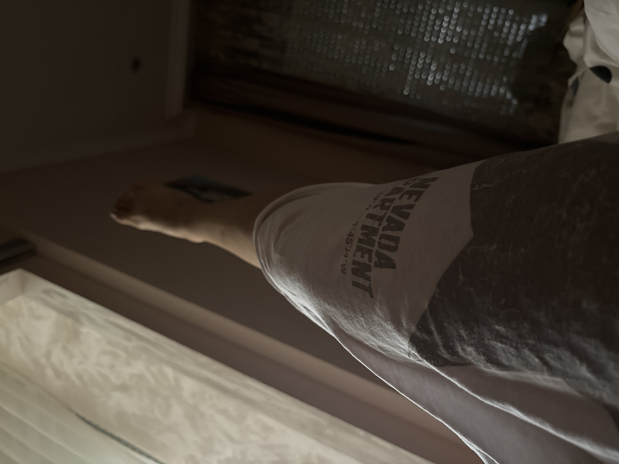 The image shows a person's leg and torso from a low angle. The person is wearing a light-colored t-shirt with a graphic print that includes the words "NEVADA DEPARTMENT". The leg is bent, with the foot visible in the upper left of the frame. The background is dimly lit, with a window covered by sheer curtains on the left and a dark, textured surface on the right. The setting appears to be indoors, possibly a bedroom, with natural light filtering through the curtains. There is no discernible information about the city of Budva, Montenegro, in the image.